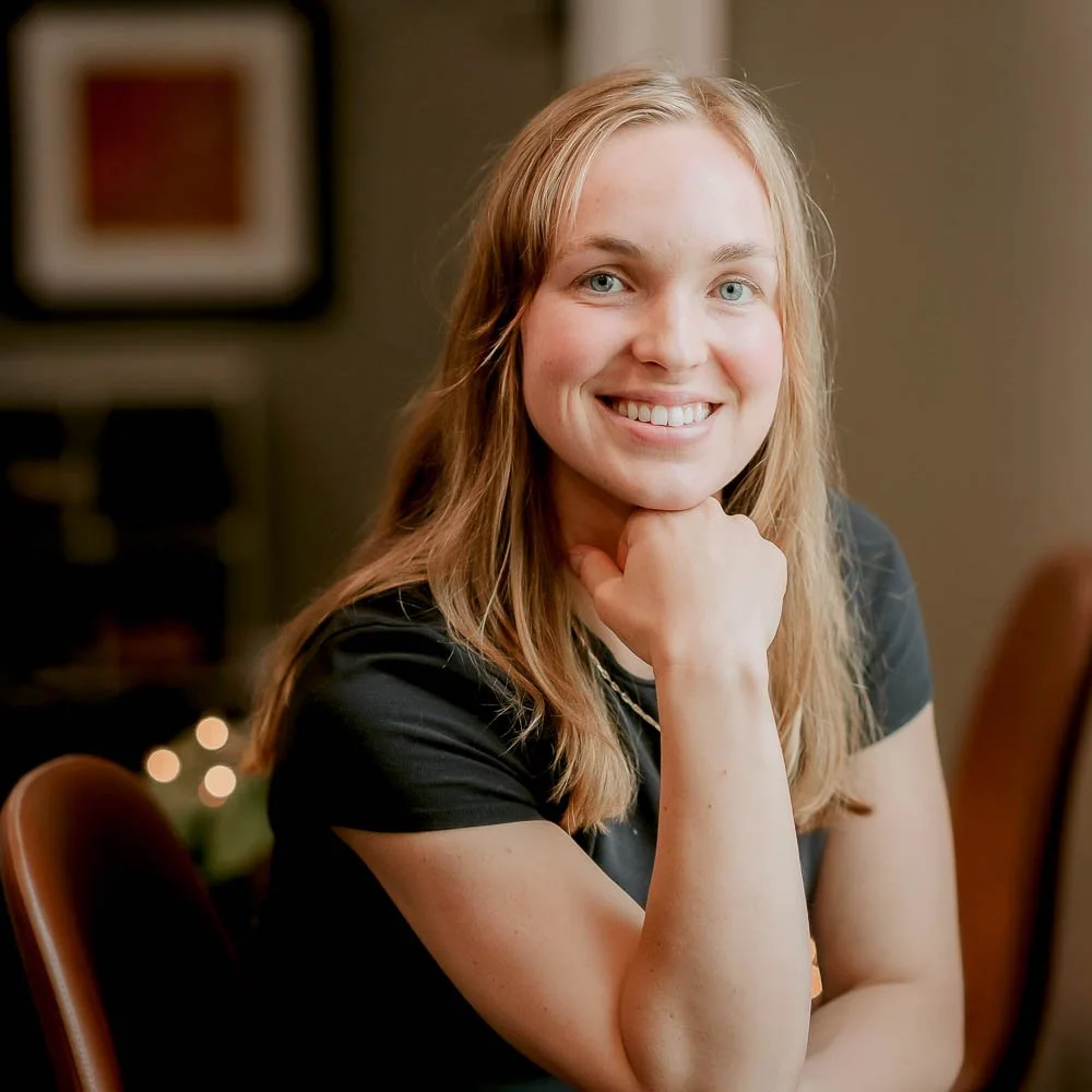 A young woman with blonde hair and blue eyes smiling and resting her chin on her hand in a warmly lit indoor setting.