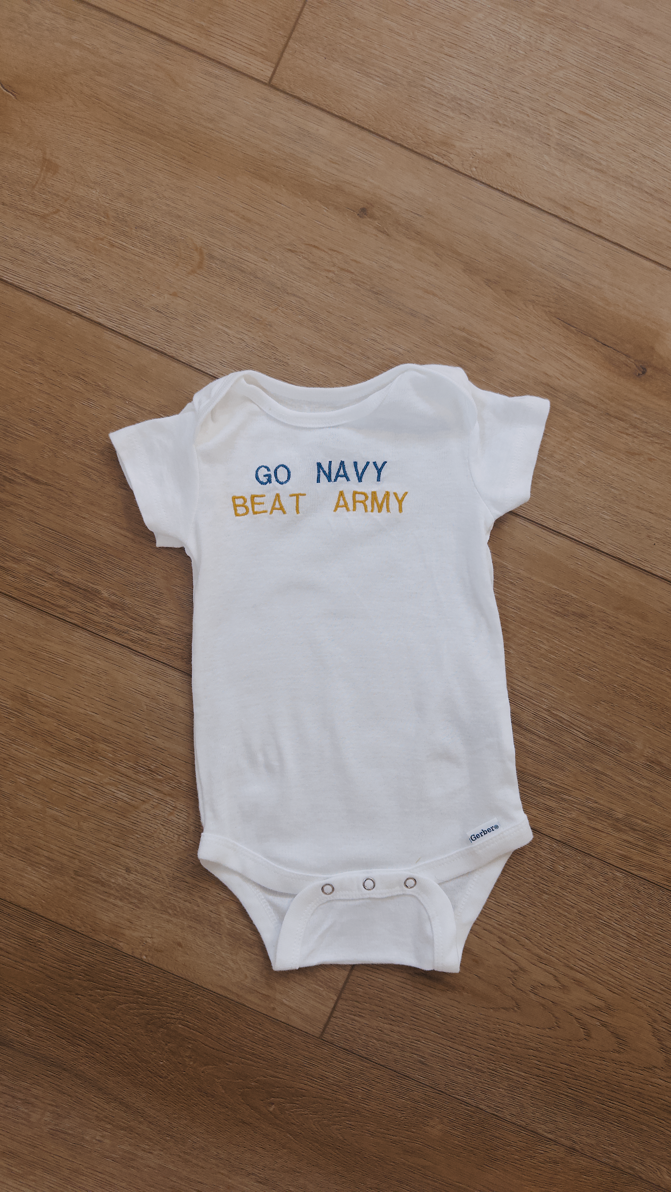 A small white baby onesie with the words "GO NAVY BEAT ARMY" embroidered in blue and yellow on the front, lying on a wooden surface.