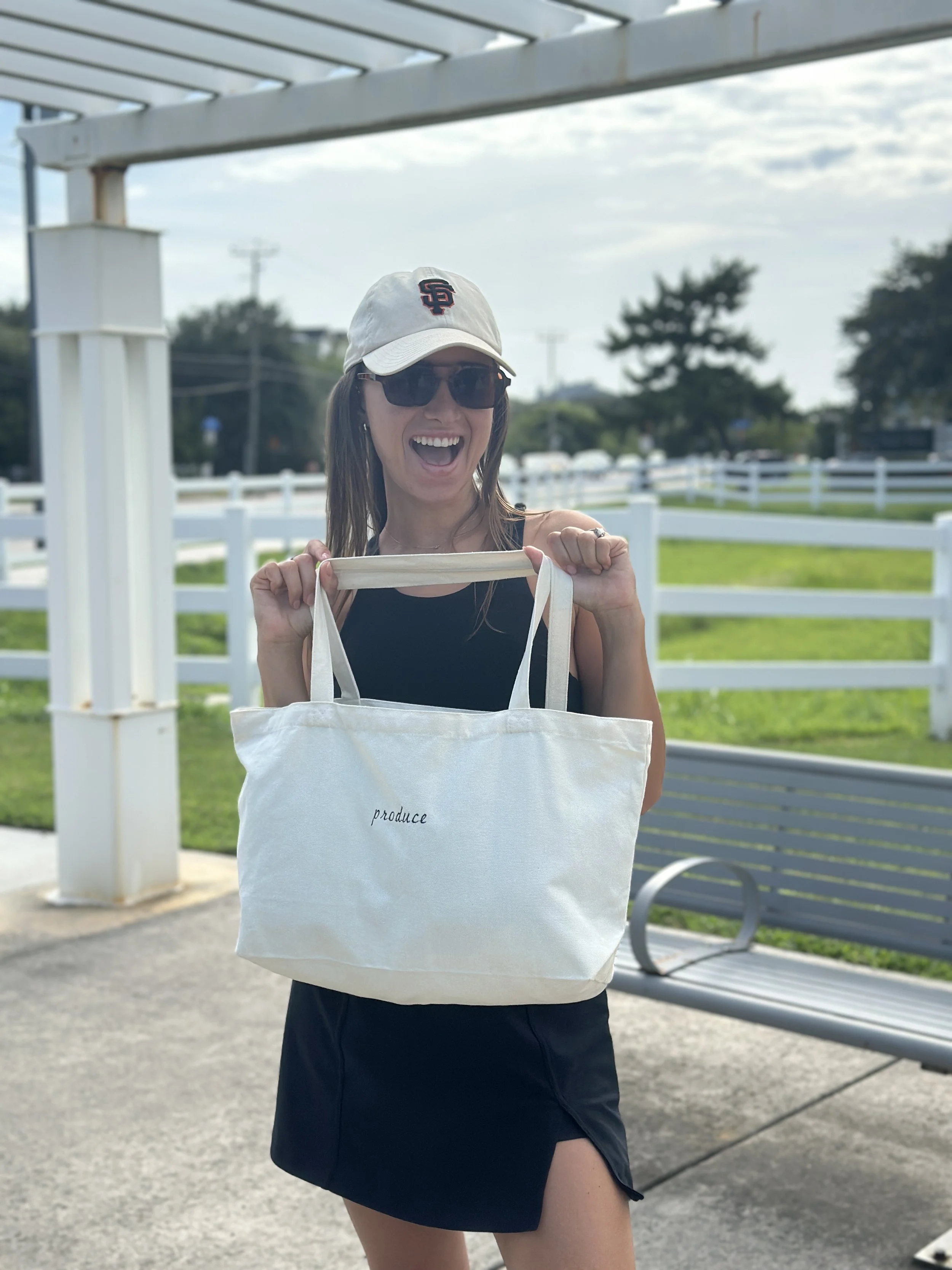 A woman holding a white tote bag that has the word 'produce' embroidered on it. She is smiling, wearing a white cap, sunglasses, a black sleeveless top, and a black skirt. She is outdoors at a train station or bus stop with white fencing, a bench, an