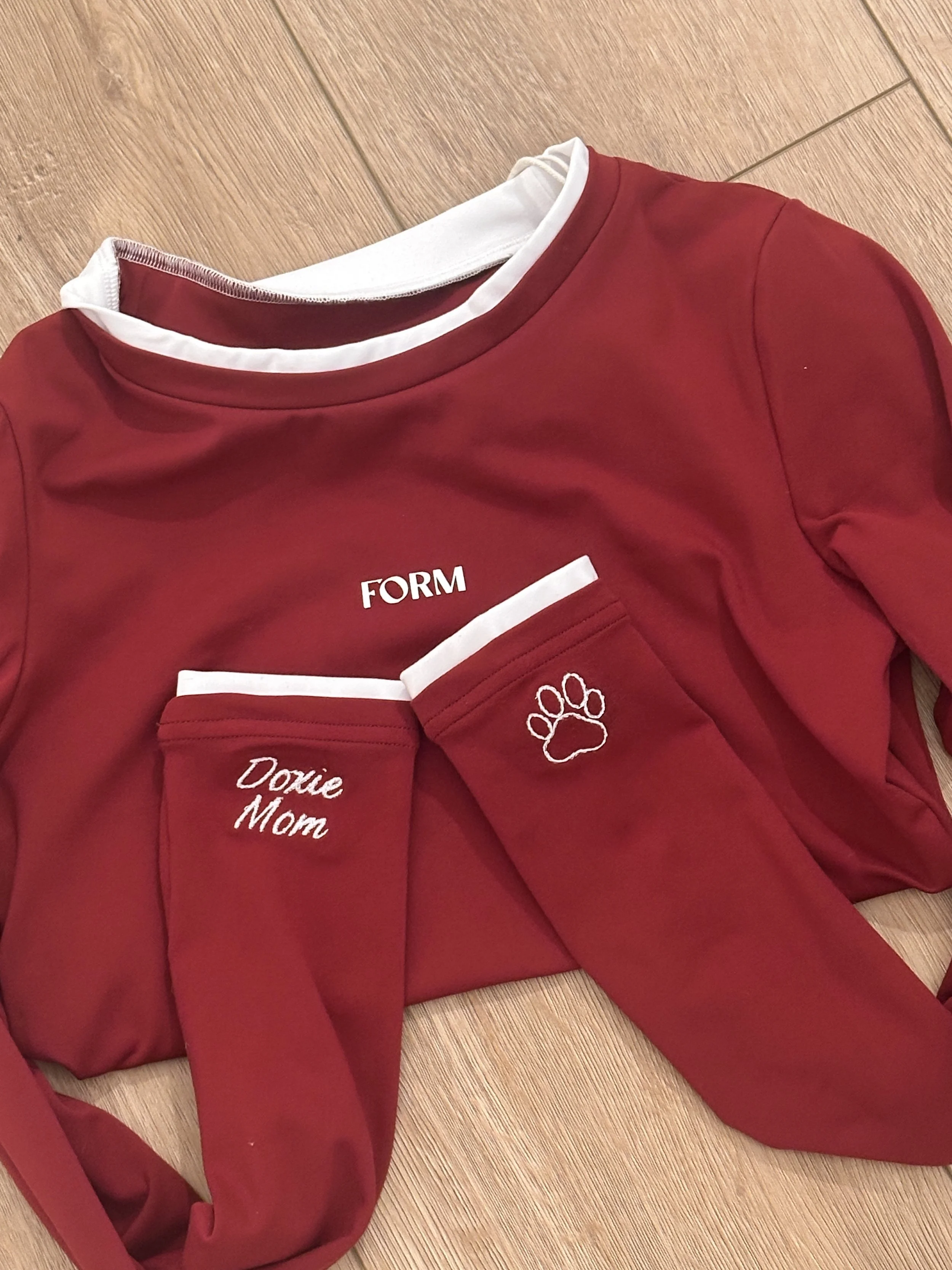 Red sweatshirt with white collar, embroidered text "FORM" on chest, and matching red sleeves with white accents; two red sleeves with embroidery "Doxie Mom" and a paw print are placed on top of the sweatshirt on a wooden floor.