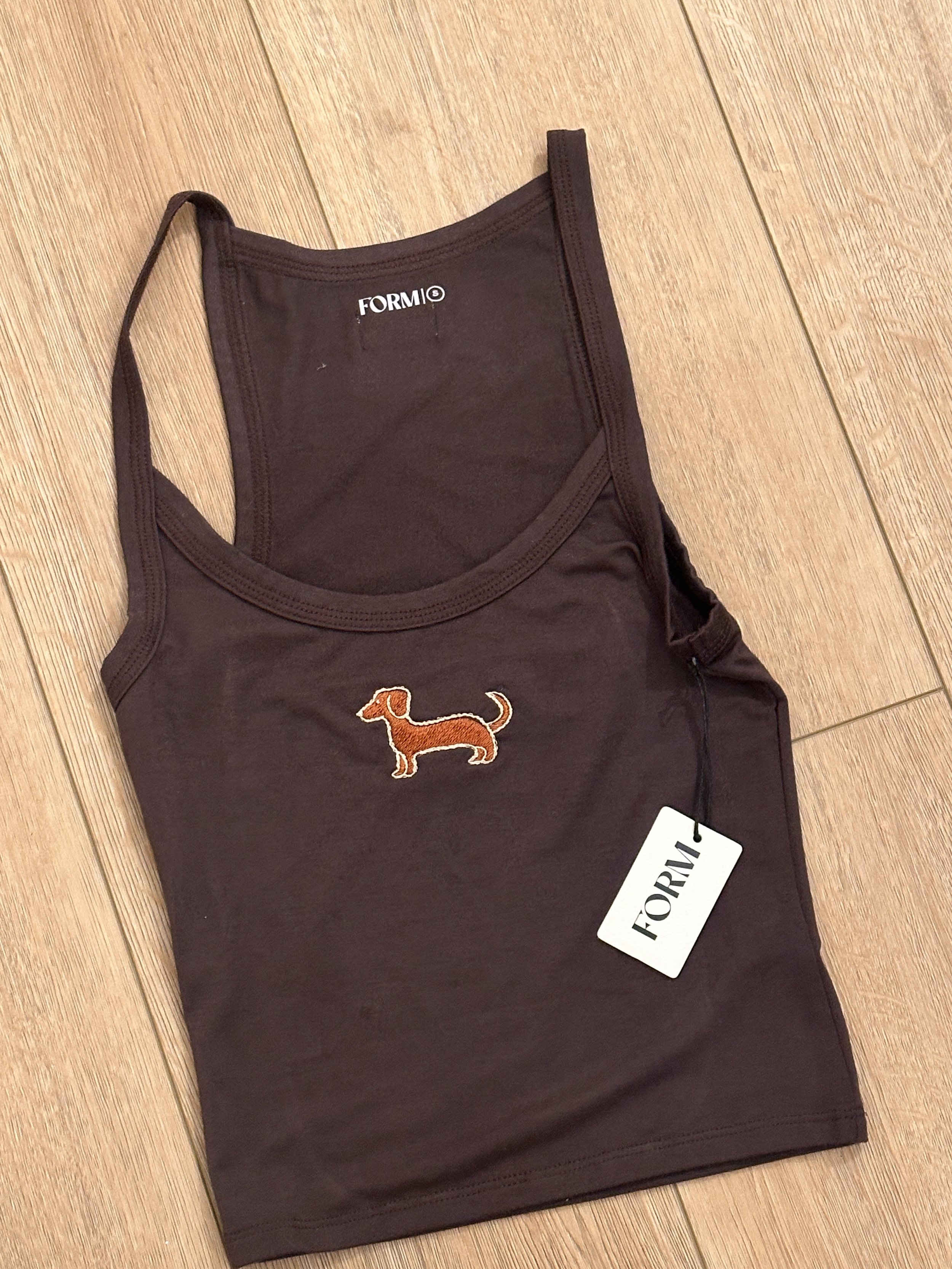 A brown tank top with a small embroidered orange dachshund on the front, laid flat on a wooden floor, with a white tag reading 'FORM' attached.