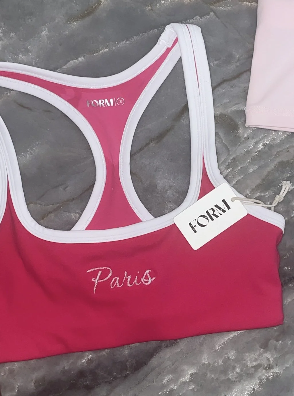 Pink sports bikini top with white trim, embroidered with 'Paris,' laid out on a marbled surface.