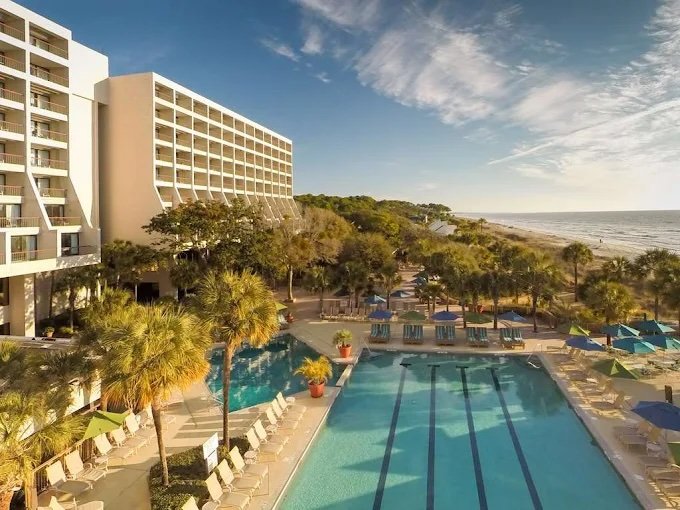 A beachfront hotel with a large outdoor pool area, surrounded by lounge chairs and umbrellas, with the ocean visible in the background under a partly cloudy sky.
