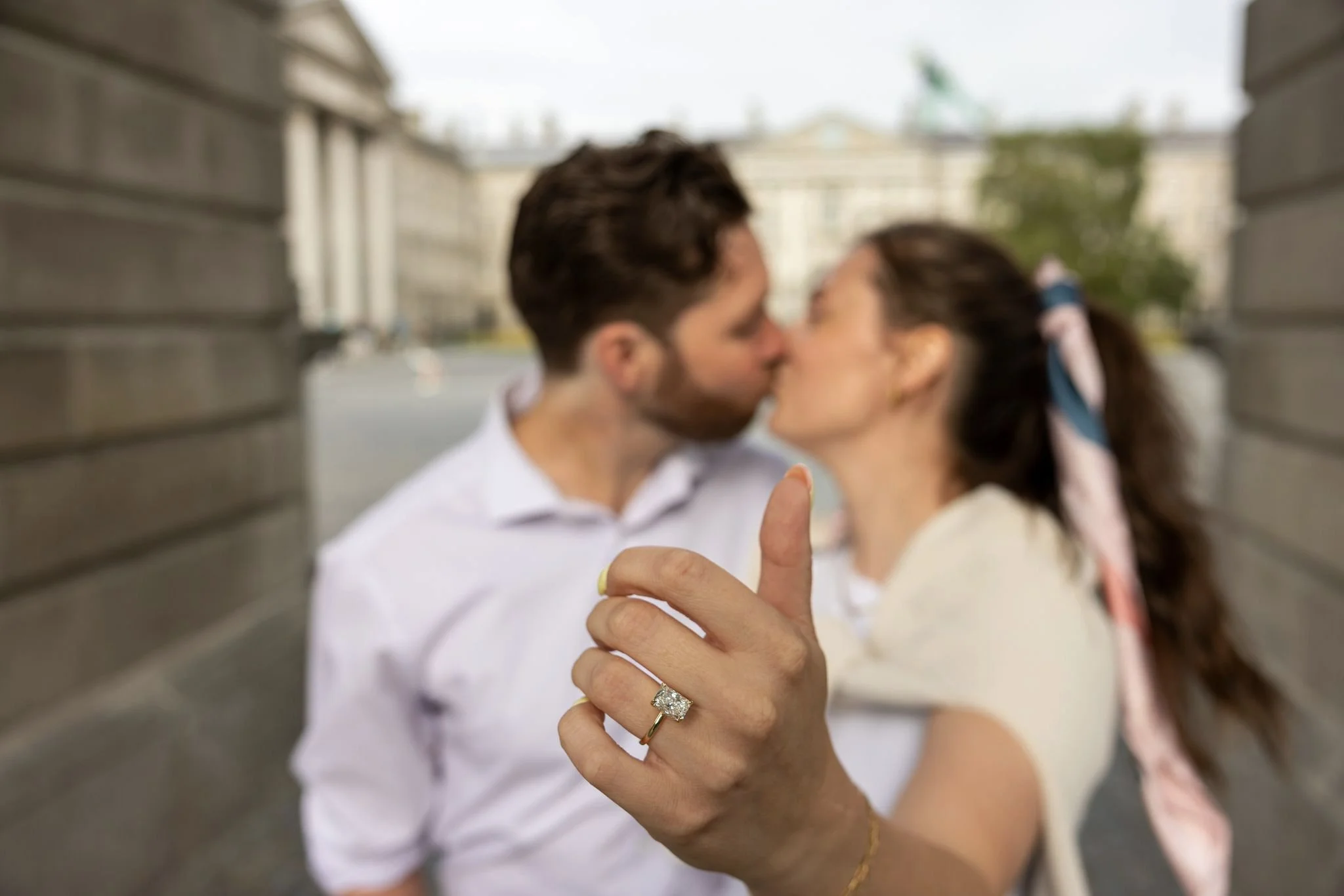 A woman showing her engagement ring while a couple kiss in the background.