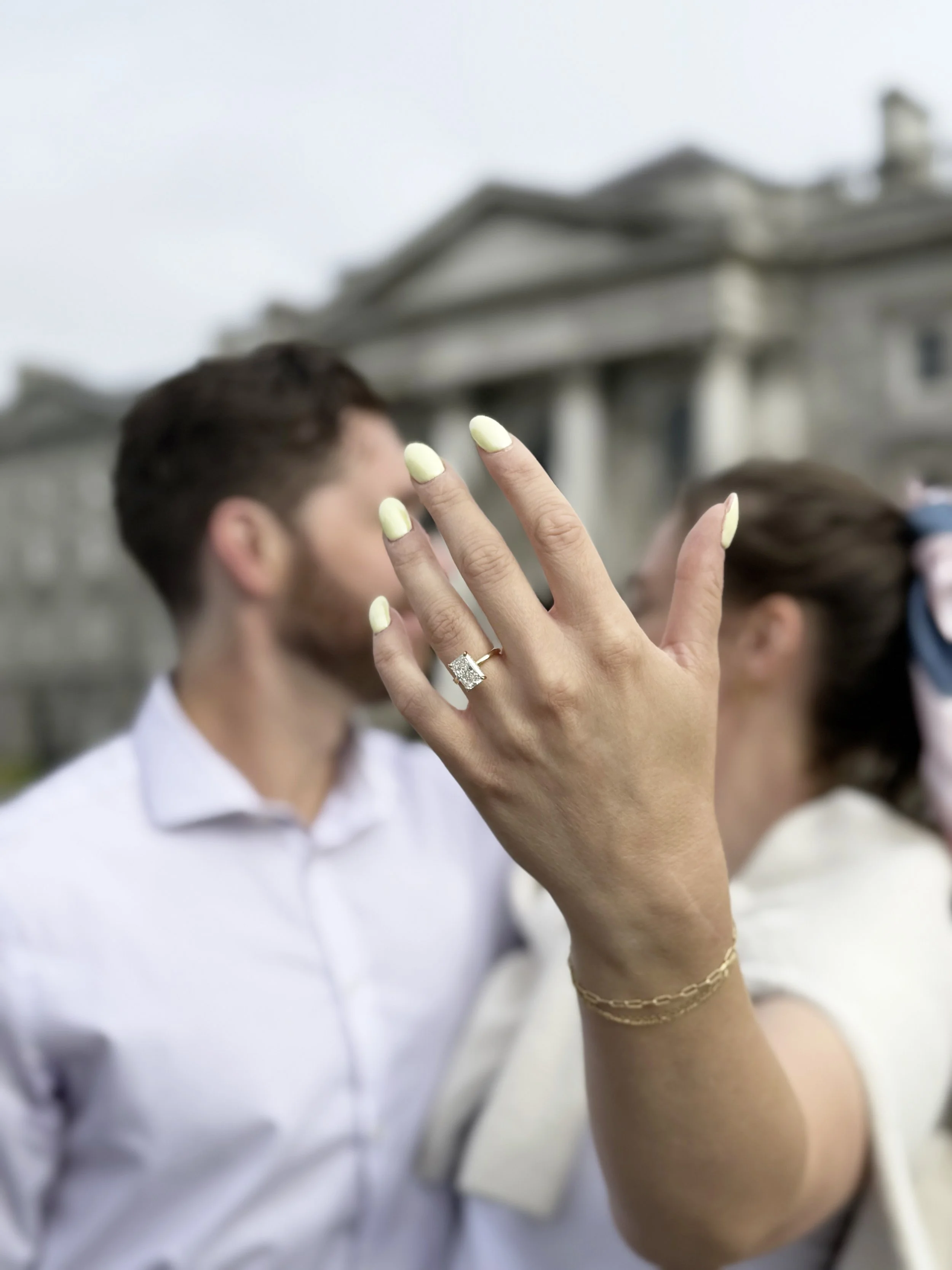 Close-up of a woman's hand showing an engagement ring, with a man and woman blurred in the background, outdoors in front of a building.