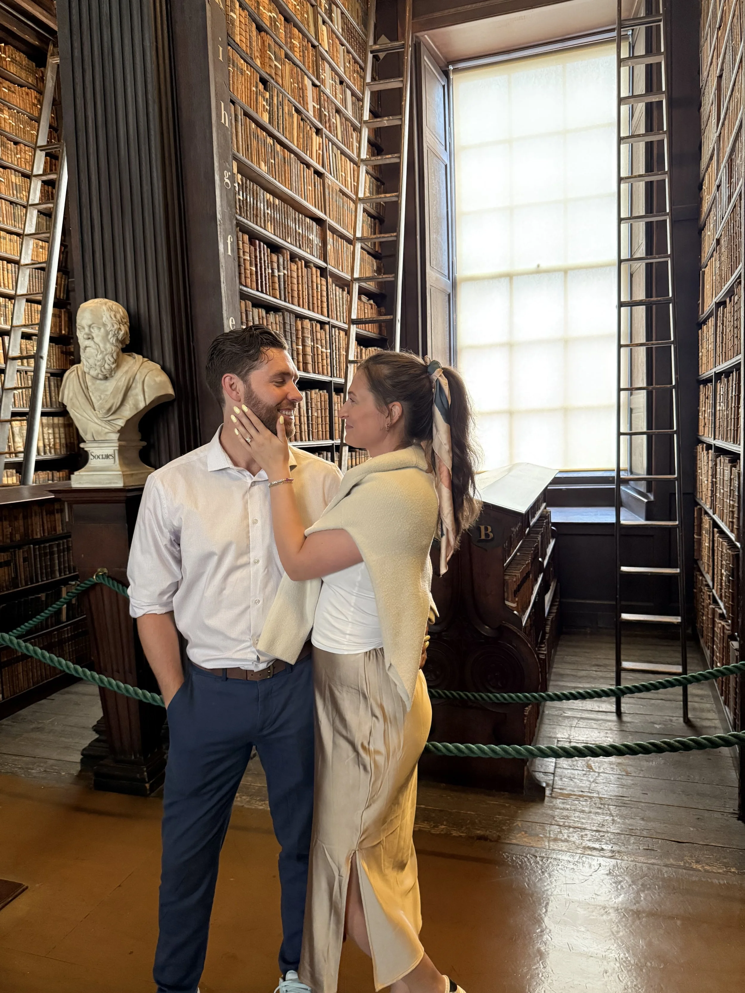 A couple standing close together in a library with tall bookshelves, a bust sculpture, ladders, and large window in the background.