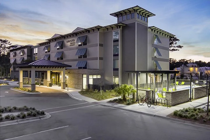 Modern multi-story hotel named 'Springhill Suites' with a parking lot and bike rack in the foreground, outdoor seating area, and a sunset sky in the background.