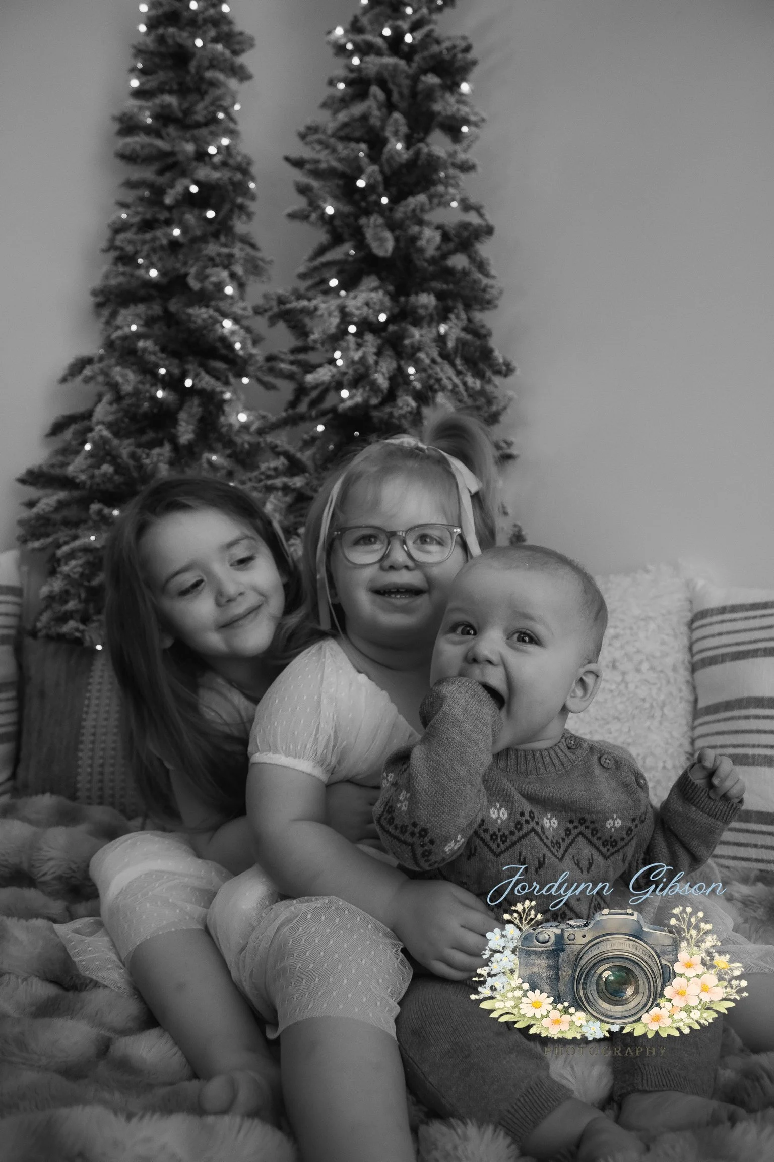 Three smiling children sitting on a cozy blanket in front of a decorated Christmas tree with lights.