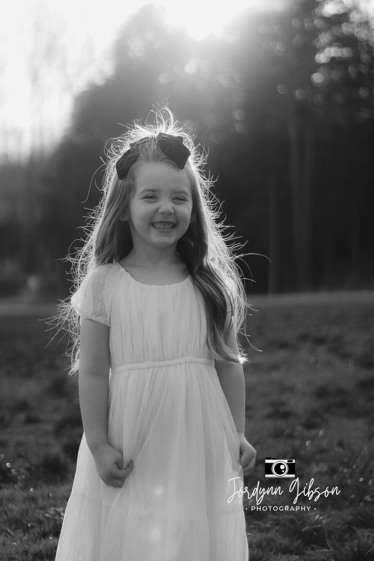 A young girl with long hair wearing a light-colored dress and a bow, smiling outdoors in a field with trees in the background, backlit by the sun.