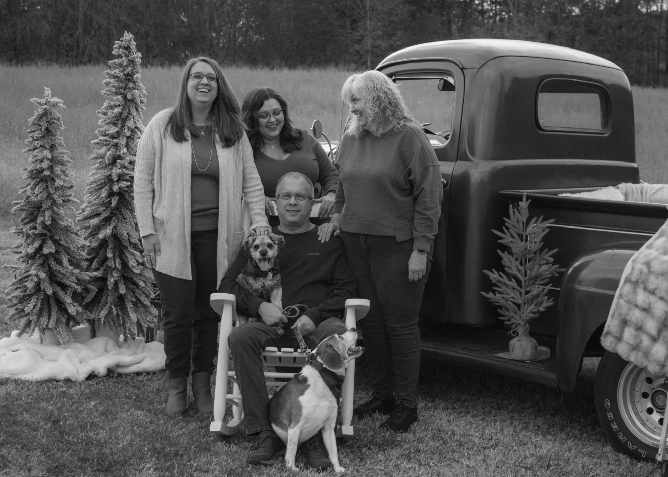 A group of five people and two dogs stand and sit outdoors in front of a vintage truck. The group includes four women and one man seated in a chair, with the women standing around him. The setting appears to be a field with some decorative Christmas 
