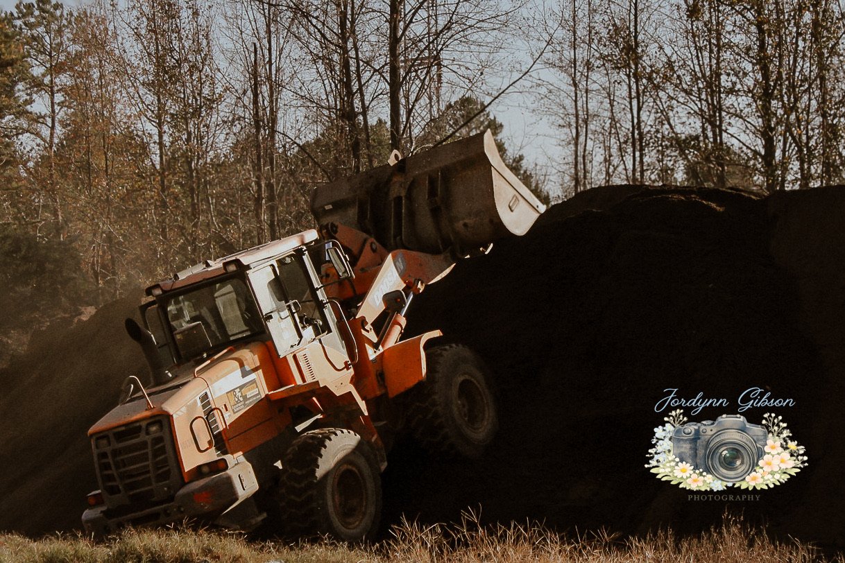 An orange front loader construction vehicle lifting a large pile of dirt or gravel on a construction site, with a background of trees and a cloudy sky.