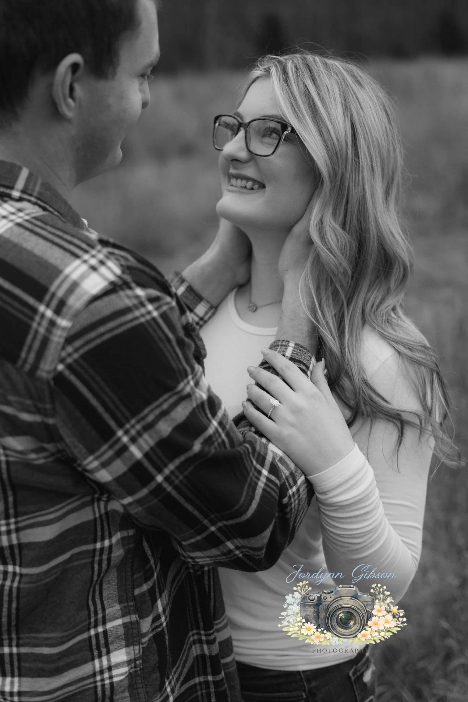 A black and white photo of a couple outdoors, with the man tenderly holding the woman's face as she smiles and looks at him.