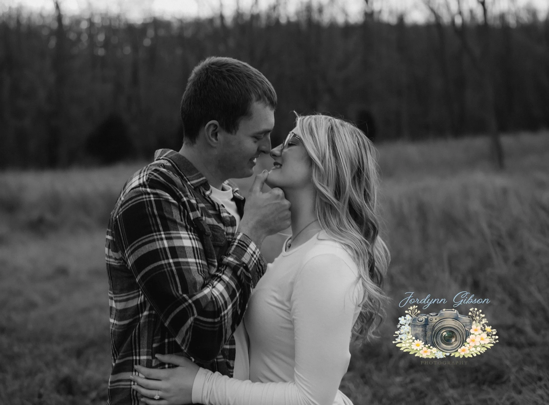 A black-and-white photo of a couple standing close in an outdoor field with trees in the background. The man gently touches the woman's chin as they smile at each other.
