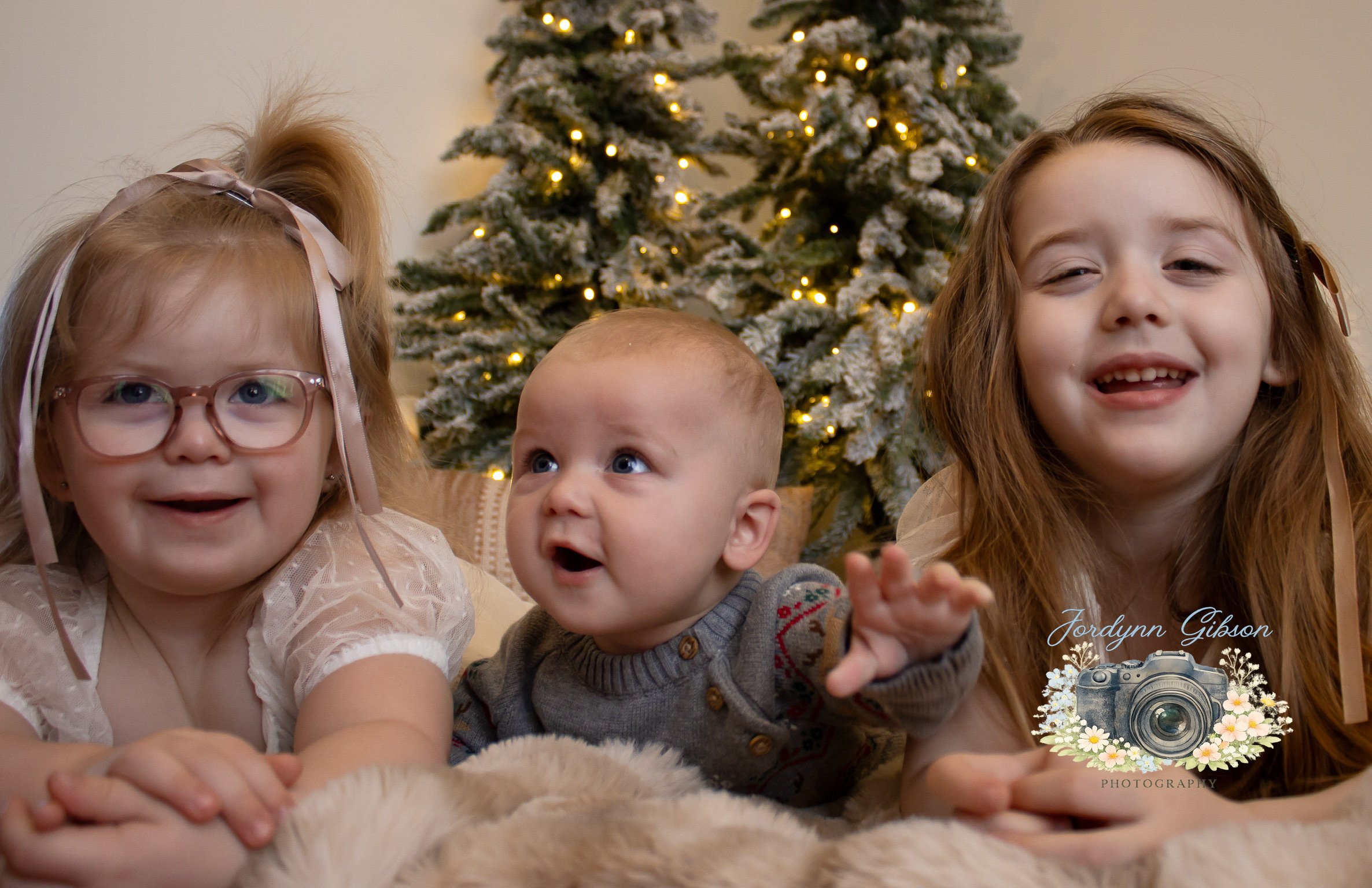 Three children with a Christmas tree in the background, lying on a cozy blanket, smiling and looking at the camera.