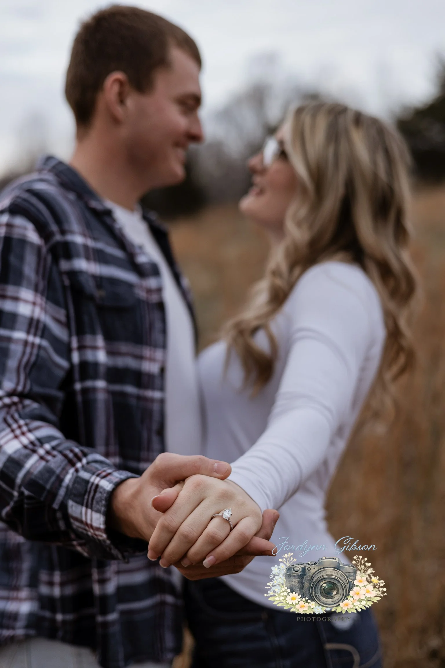 A couple standing outdoors, holding hands and smiling at each other. The woman is wearing a white long-sleeve top and has an engagement ring on her finger. The man is wearing a plaid shirt.