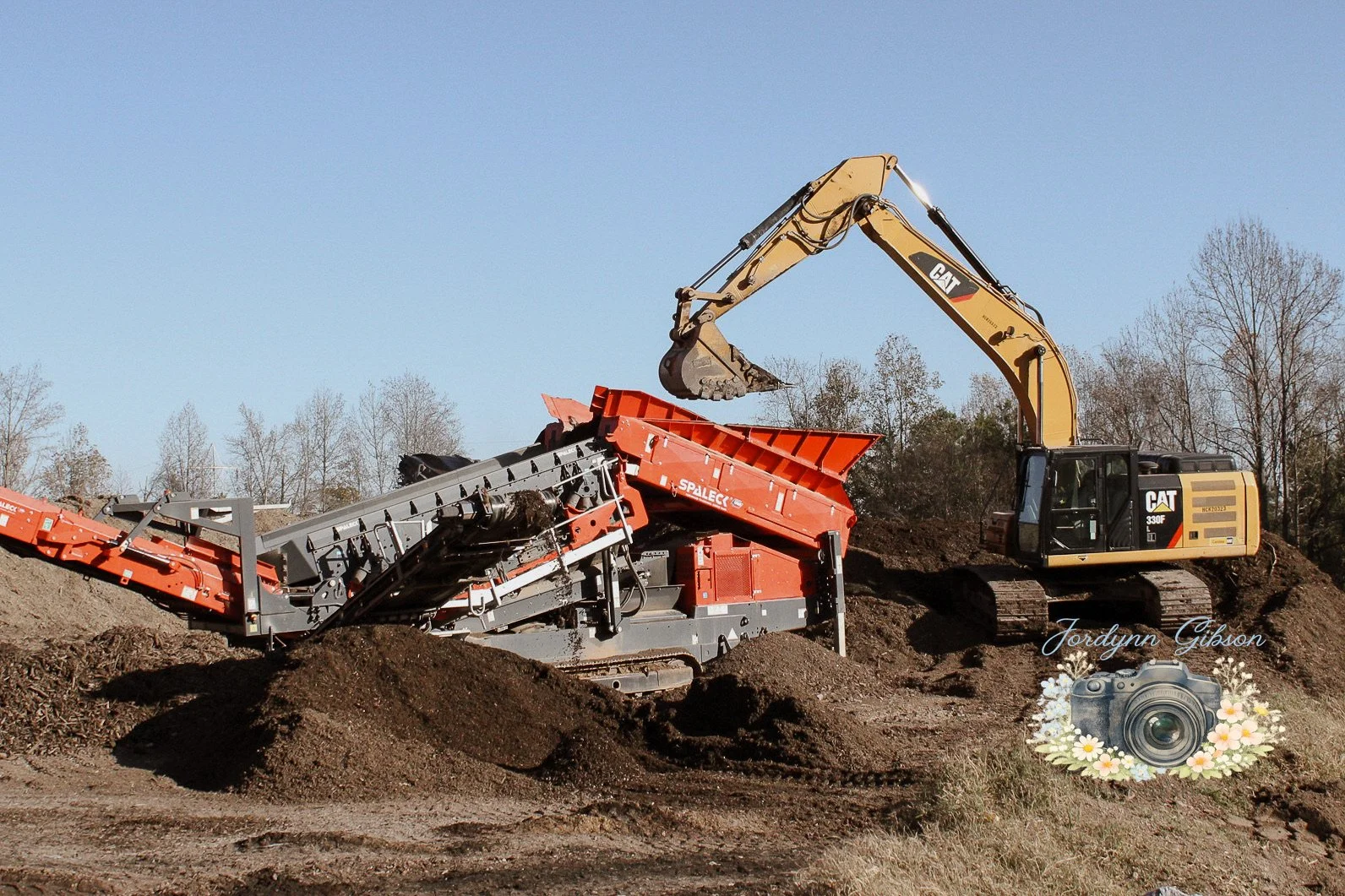 A construction scene with an excavator loading dirt onto a conveyor belt in an outdoor area with trees in the background.