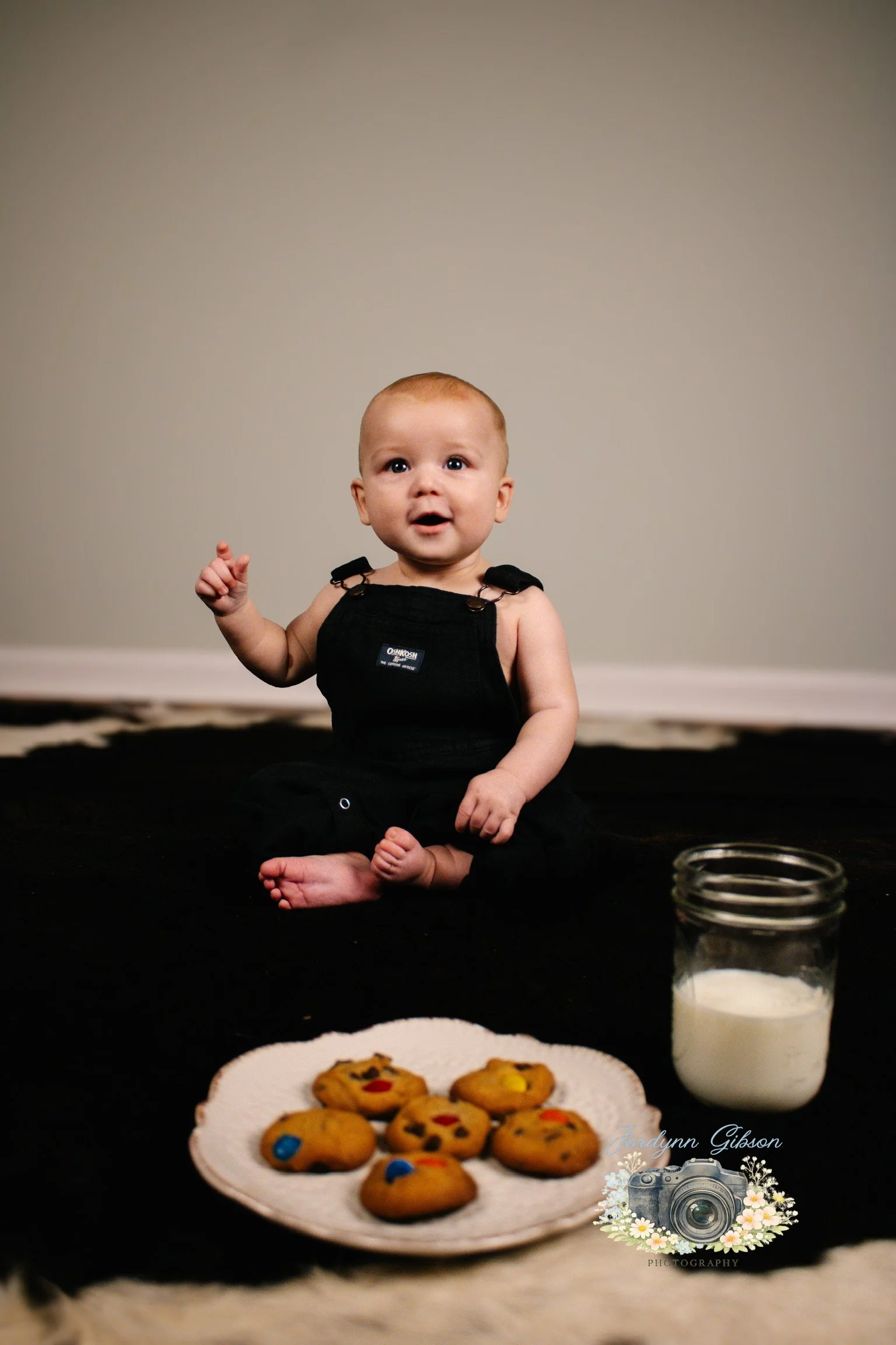 A young baby in black overalls sits on a black surface with a plate of decorated cookies, a glass jar of milk, and a camera logo watermark in the foreground. The background is a plain light-colored wall.
