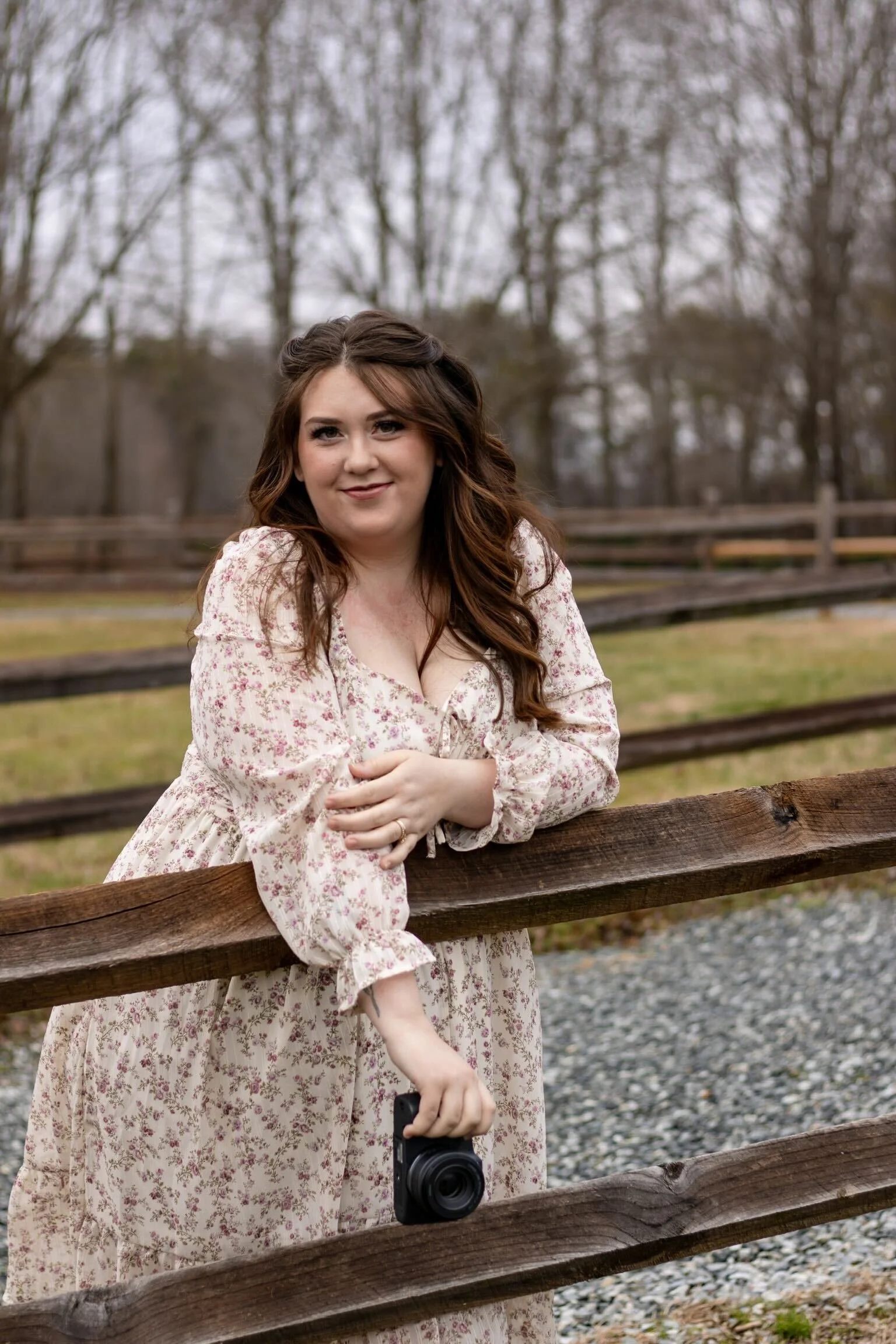 A woman with long brown hair wearing a floral dress, standing outdoors near a wooden fence, holding a camera in her right hand, with trees and overcast sky in the background.