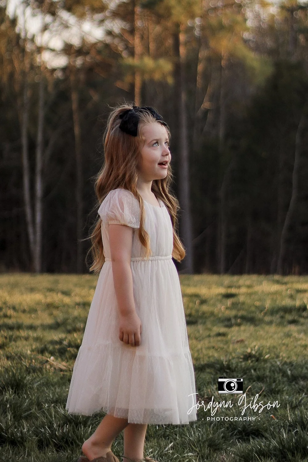 Young girl in a white dress standing on a grassy field with trees in the background, looking upward with an expression of wonder.