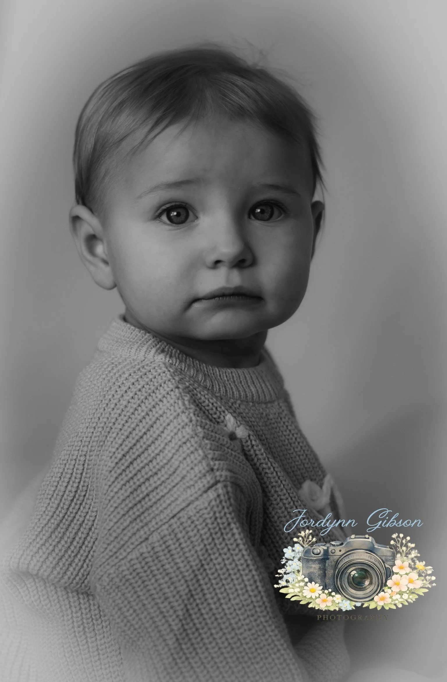 A black and white portrait of a young child with short hair, wearing a knitted sweater, looking directly at the camera with a serious expression.