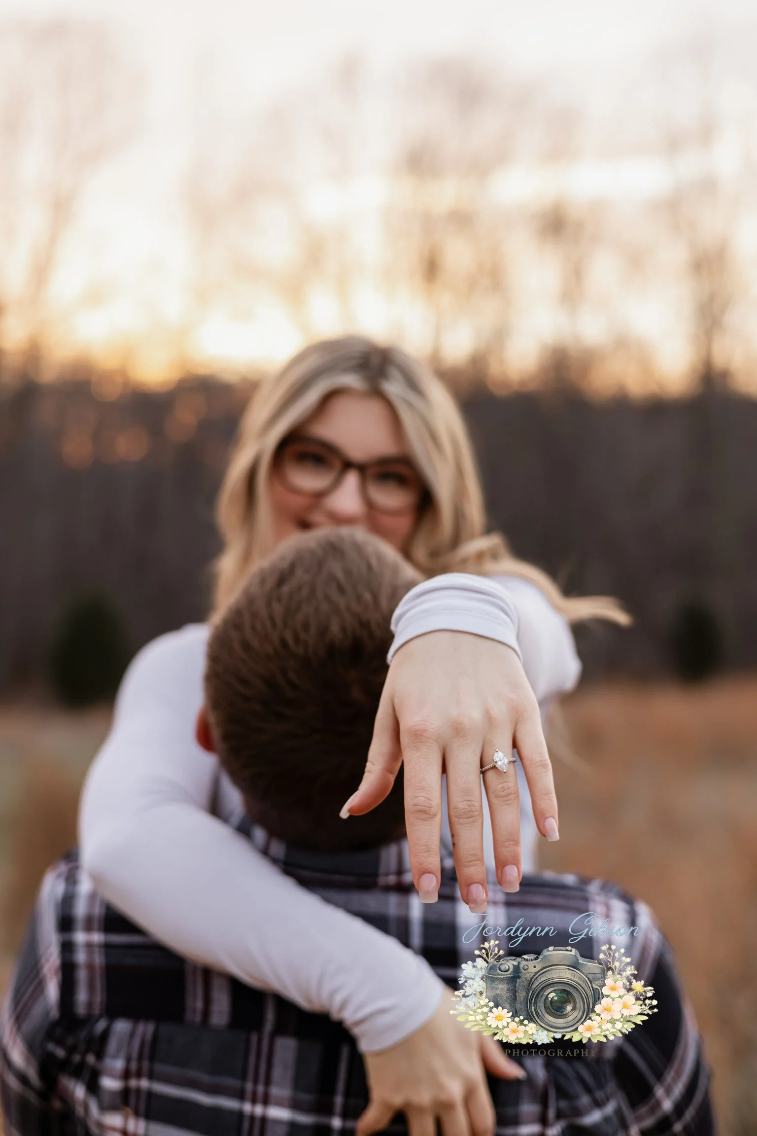 A woman with blond hair and glasses smiling while embracing a man from behind outdoors during sunset. The woman displays a ring on her left hand.