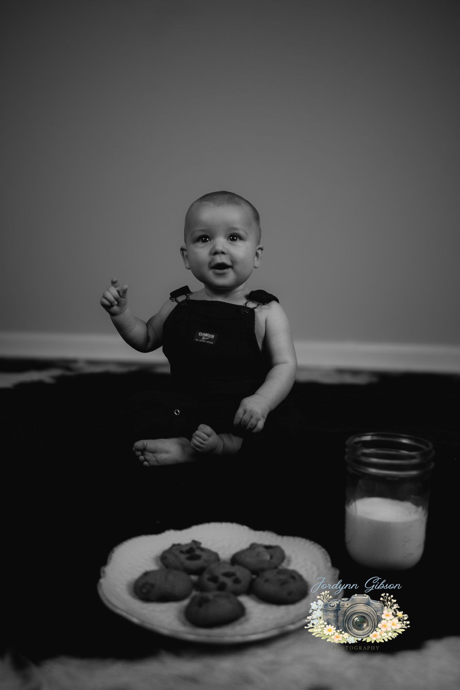 Black and white photo of a baby sitting on a blanket with a plate of cookies and a jar of milk in front of them.