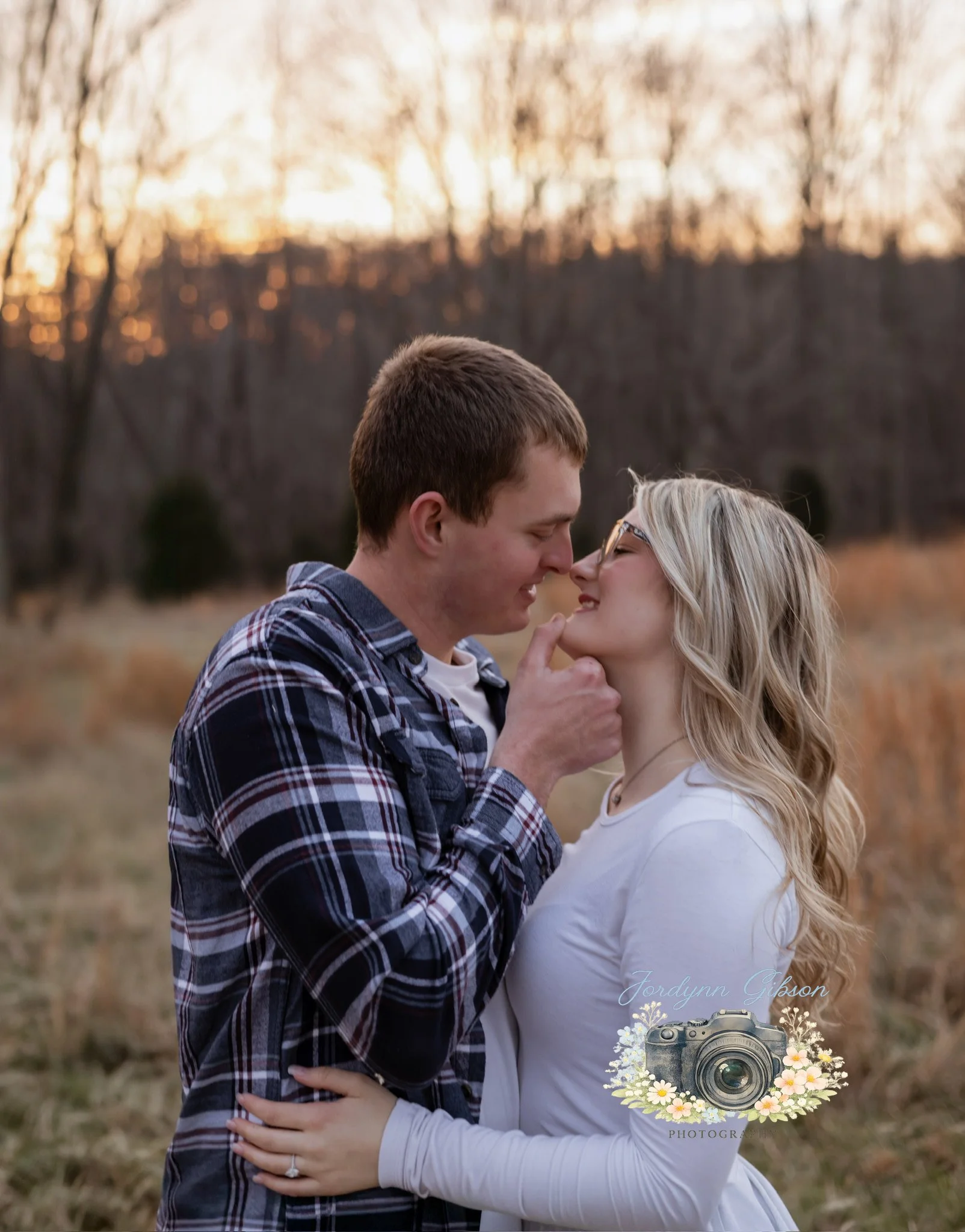 A couple stands close together in an outdoor field at sunset, about to kiss, with the woman's hand on the man's waist and their noses touching.