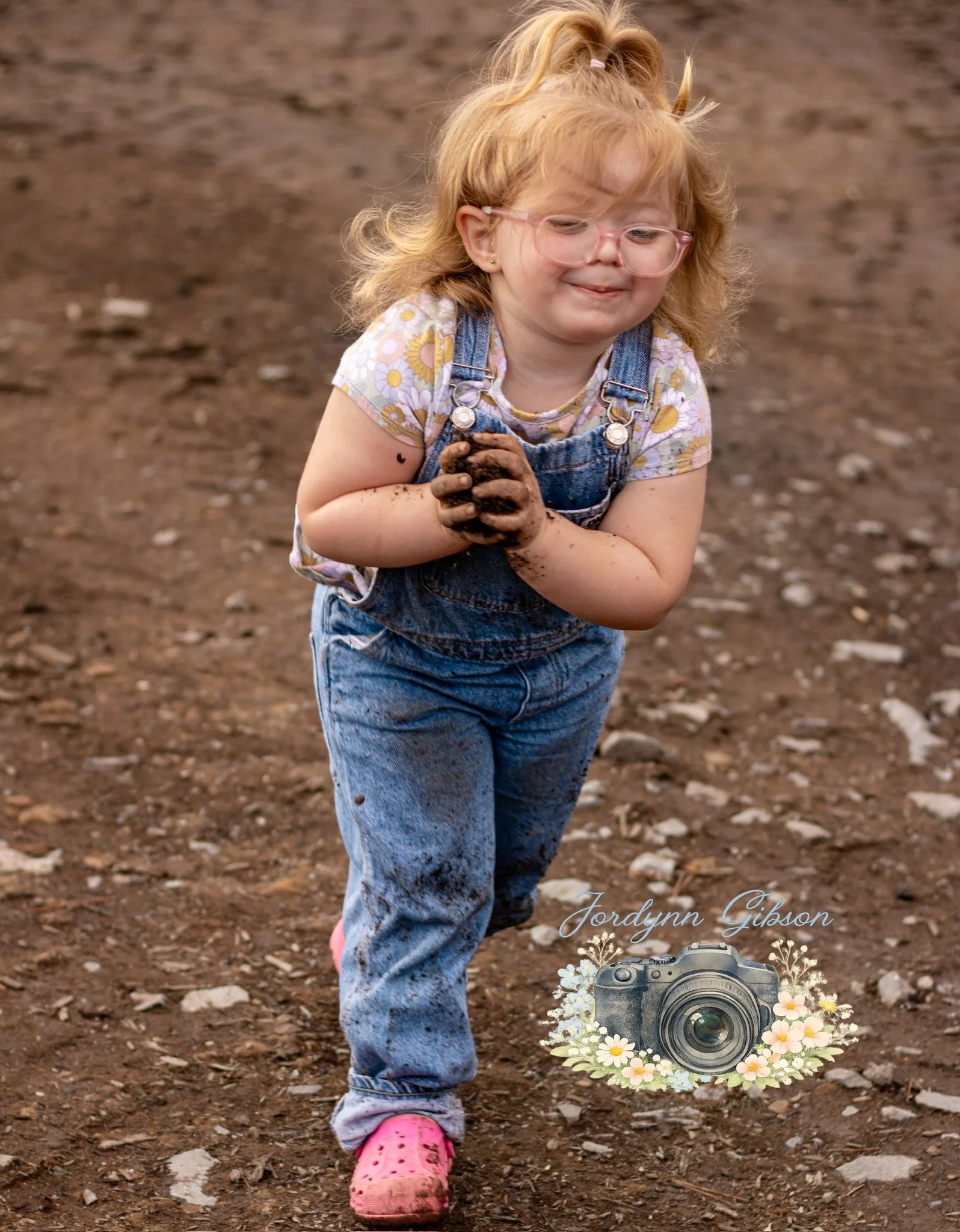 A young girl with glasses, red hair tied up, and mud on her hands and jeans walking on dirt ground, smiling.