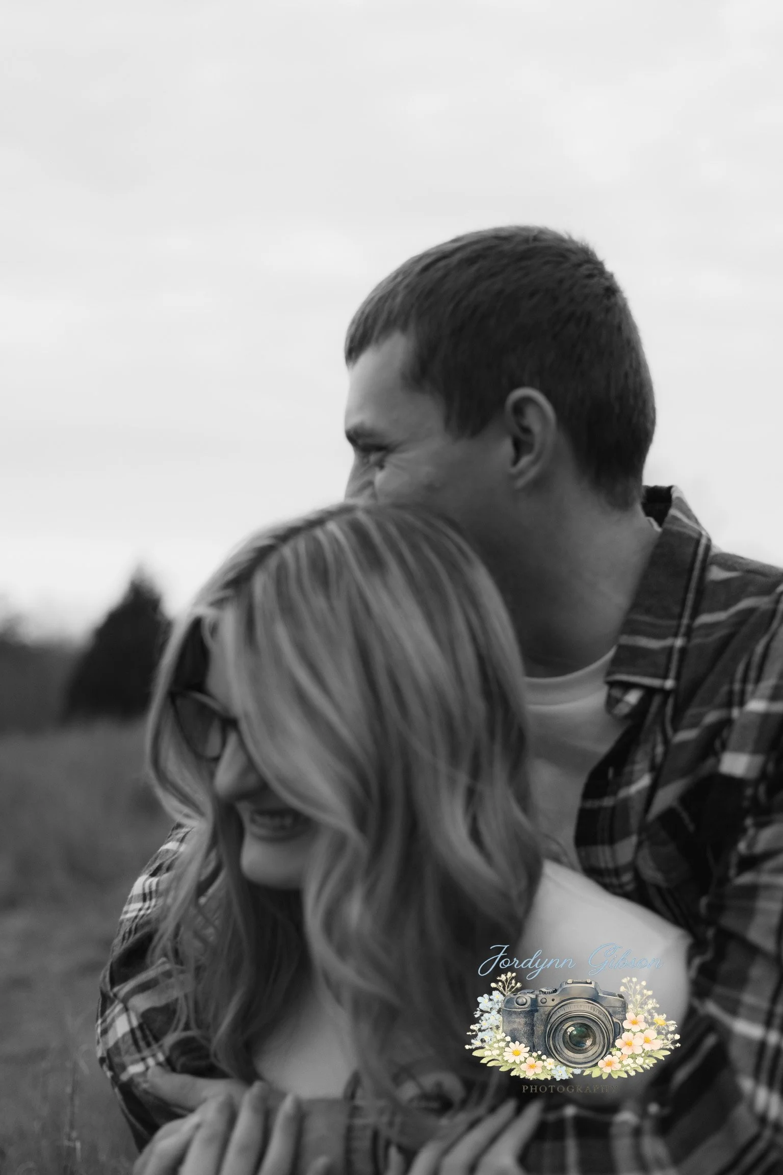 A black and white photo of a man and woman embracing outdoors, with a campfire in the background, and a logo with a camera and flowers reading 'Jordynn Gibson Photography' in the bottom corner.