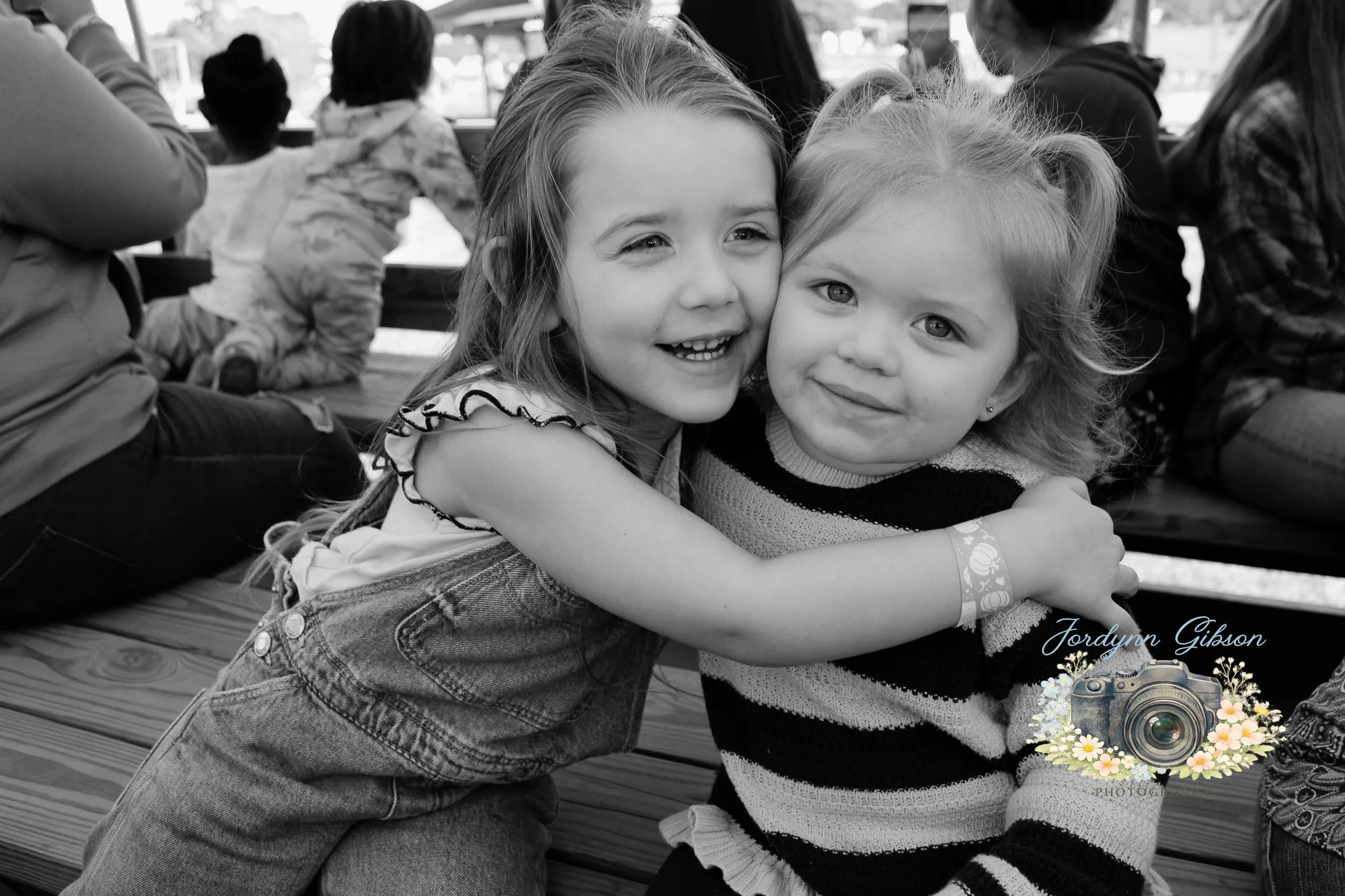 Two young girls hugging and smiling at the camera, sitting at an outdoor event with people in the background.