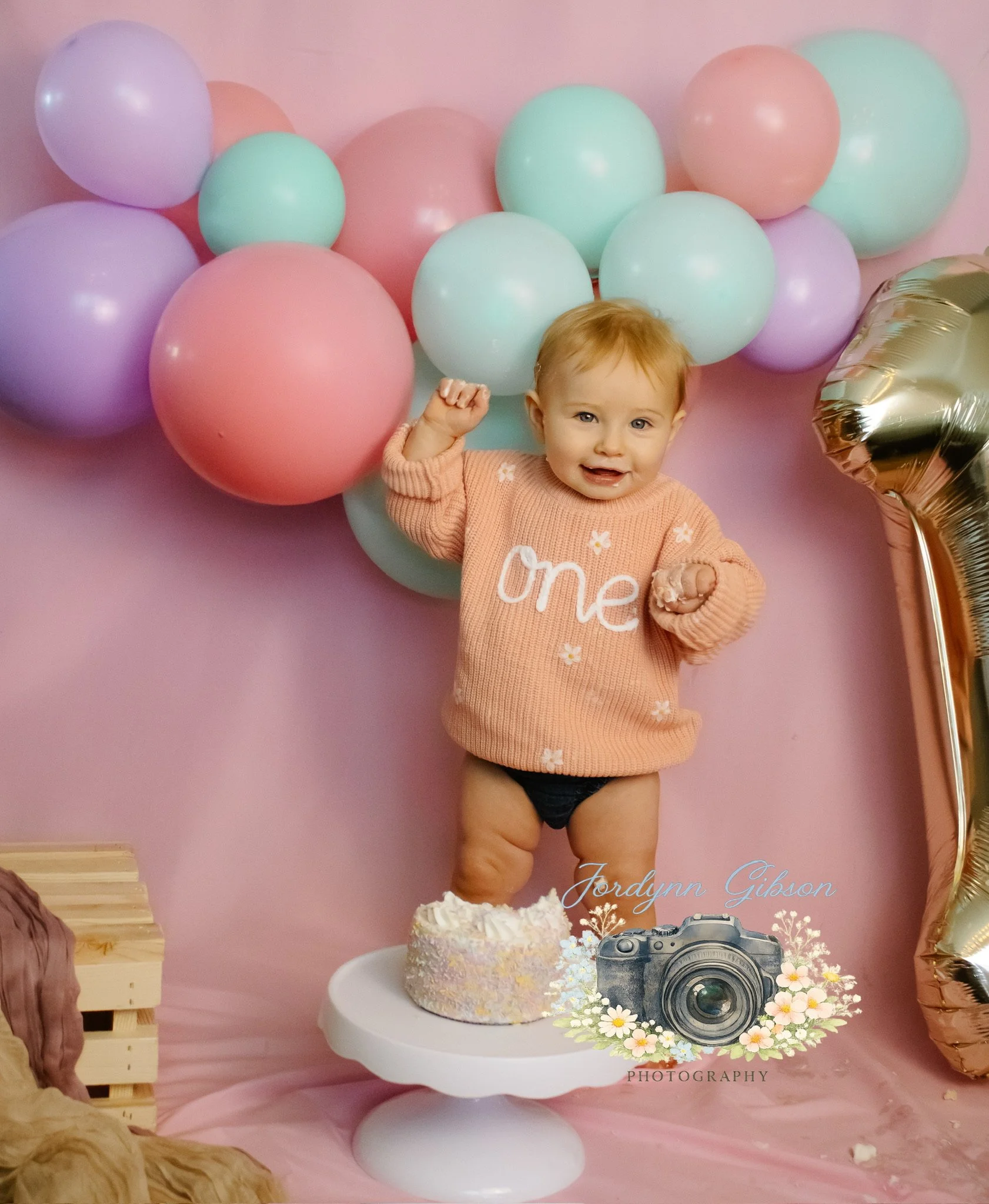 A young child celebrating their first birthday standing on a cake stand with a small cake, surrounded by pastel colored balloons, wearing a pink sweater with 'one' written on it and no pants.