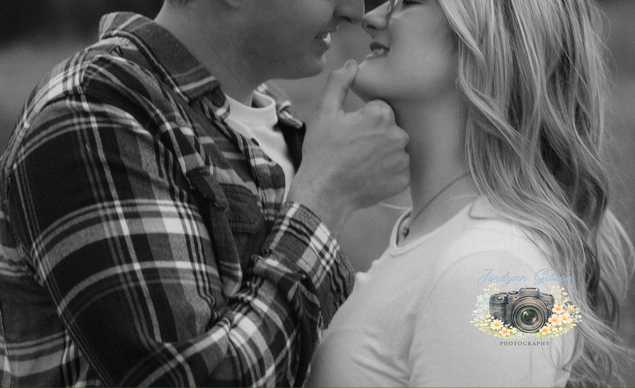 A black and white photo of a couple close together, about to kiss. The man is wearing a plaid shirt and the woman has long, wavy hair. The image features a logo with a camera and floral design, with the text "Jordynn Gibson Photography."