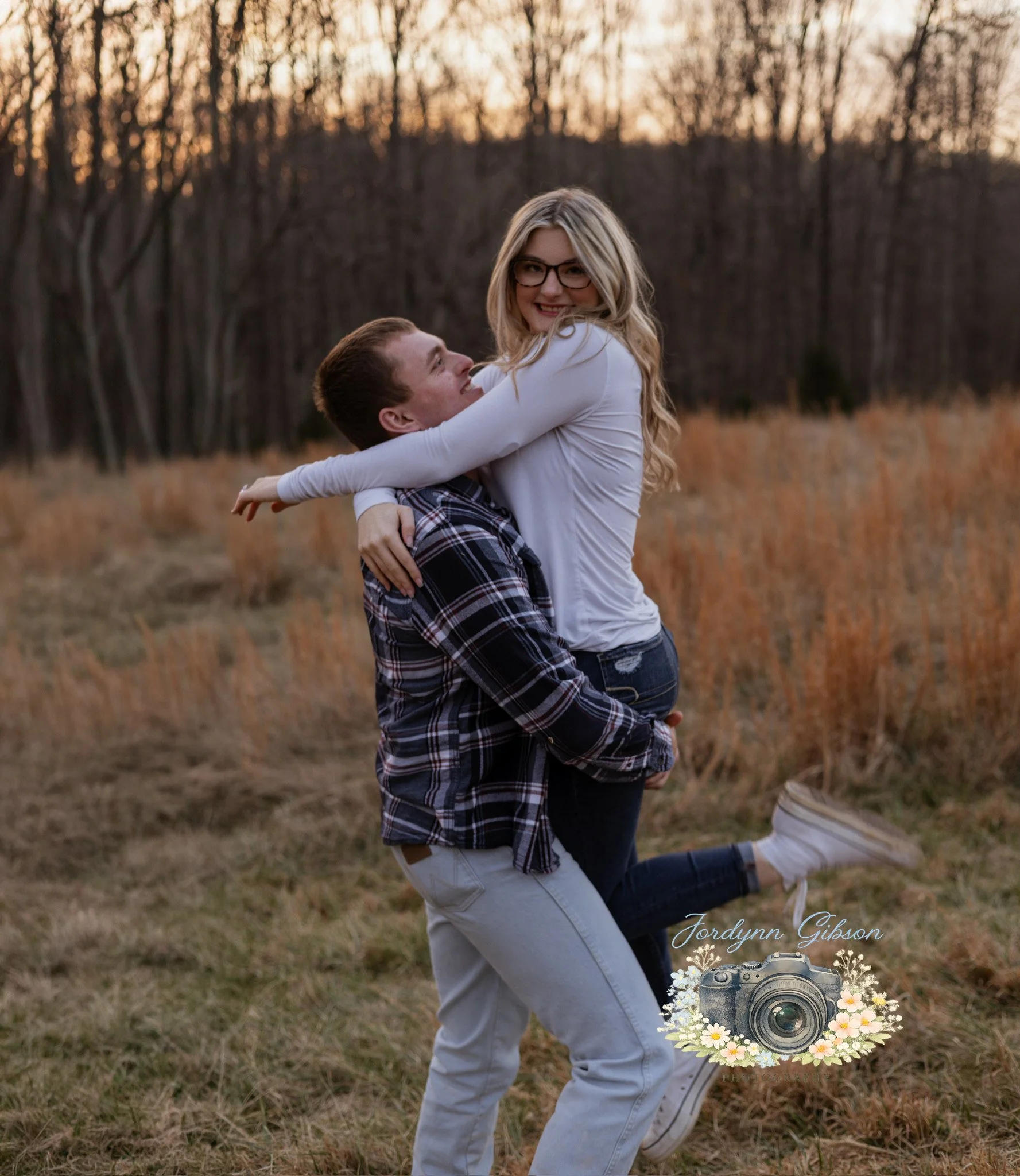 A young couple enjoying a playful moment outdoors during sunset; the man is holding the woman up, and they are smiling at each other in a field with trees in the background.