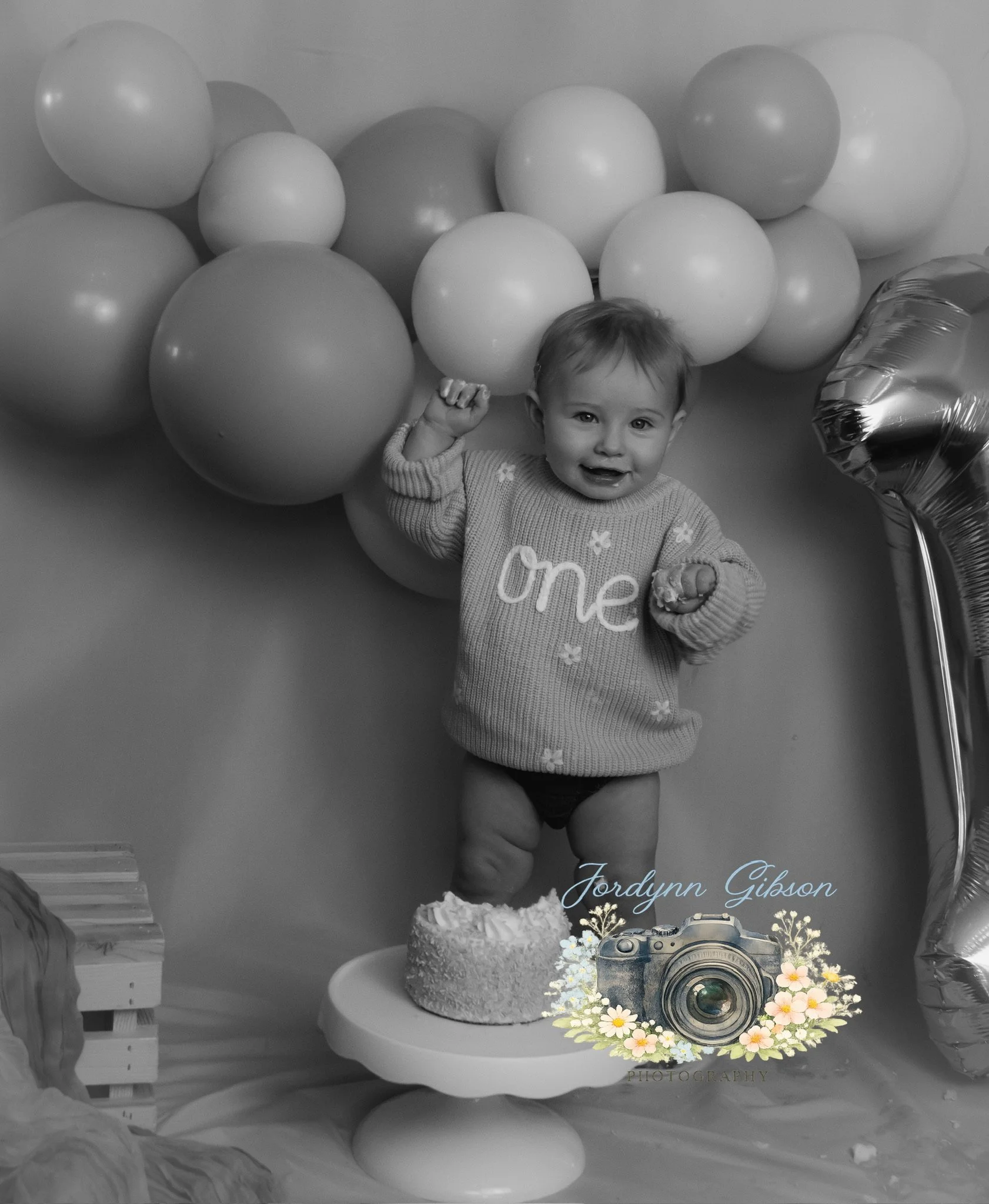 A smiling baby standing on a cake stand with a small cake, wearing a sweater that says 'one', surrounded by balloons and a large number one foil balloon.