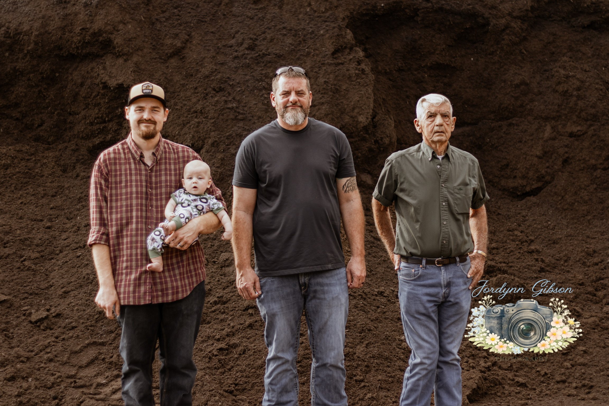Four men and a baby standing in front of a large mound of dirt. The man on the left is holding a baby, and the others are standing with hands in pockets. The background is a dirt mound.