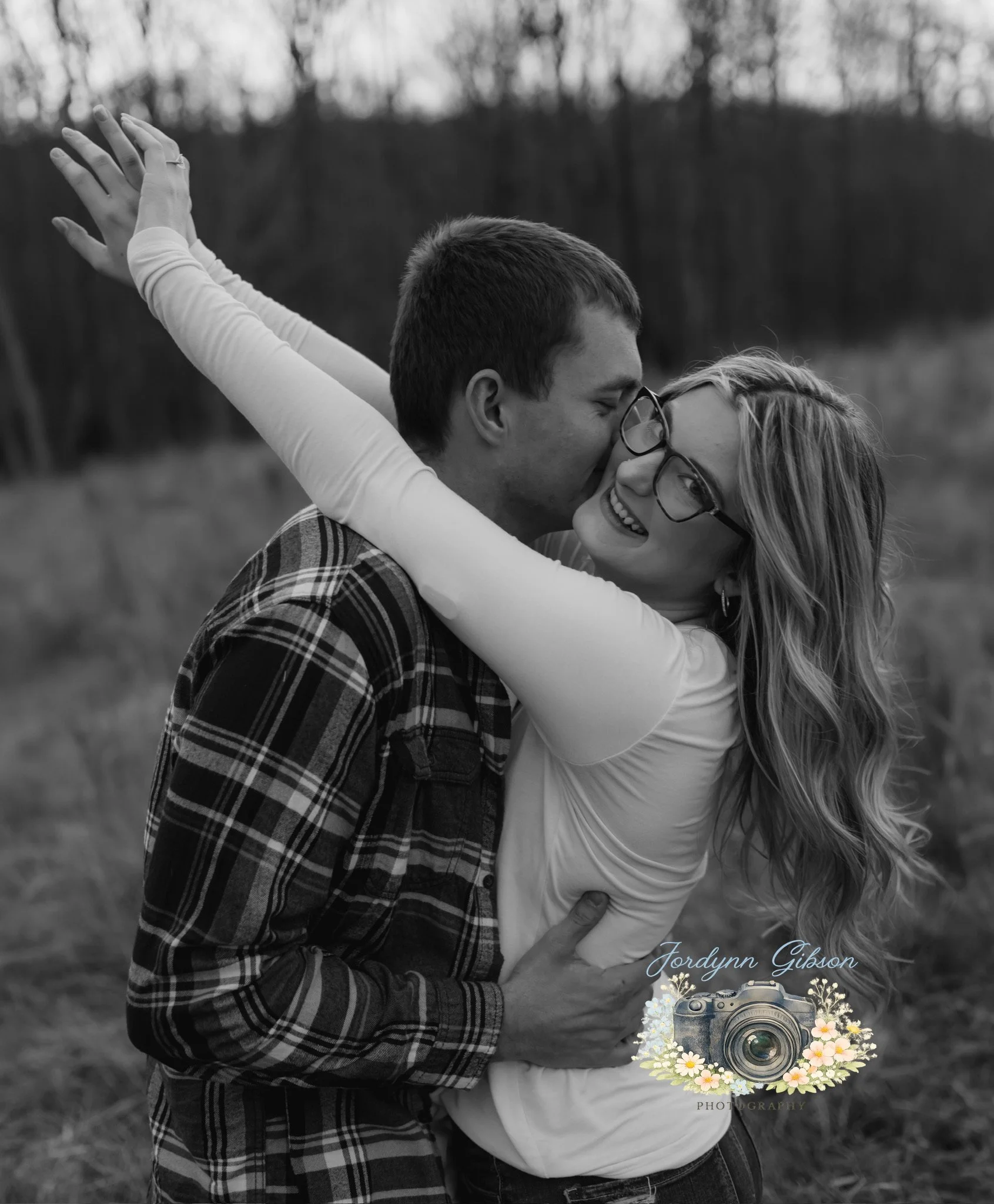 A black-and-white photo of a happy couple outdoors, with the man kissing the woman on the cheek as she smiles with glasses, her arms raised around his neck.