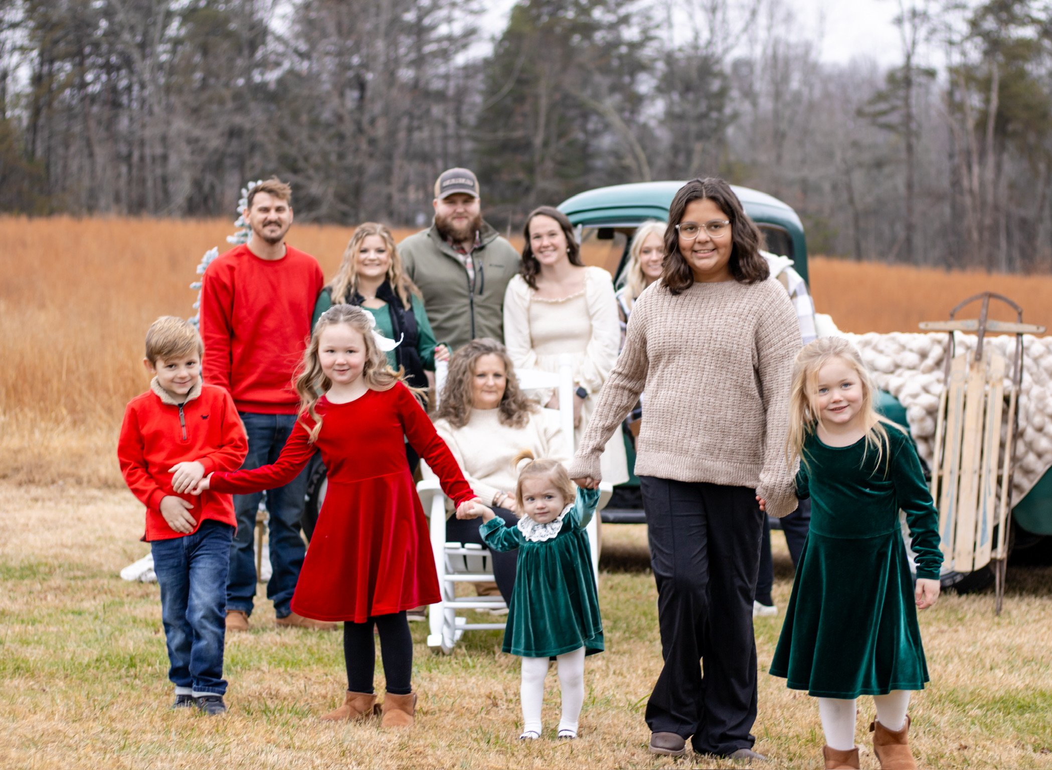 A large family gathering outdoors during fall, with people of various ages holding hands and smiling, a vintage vehicle in the background loaded with pinecones, set in an open field with trees and cloudy skies.