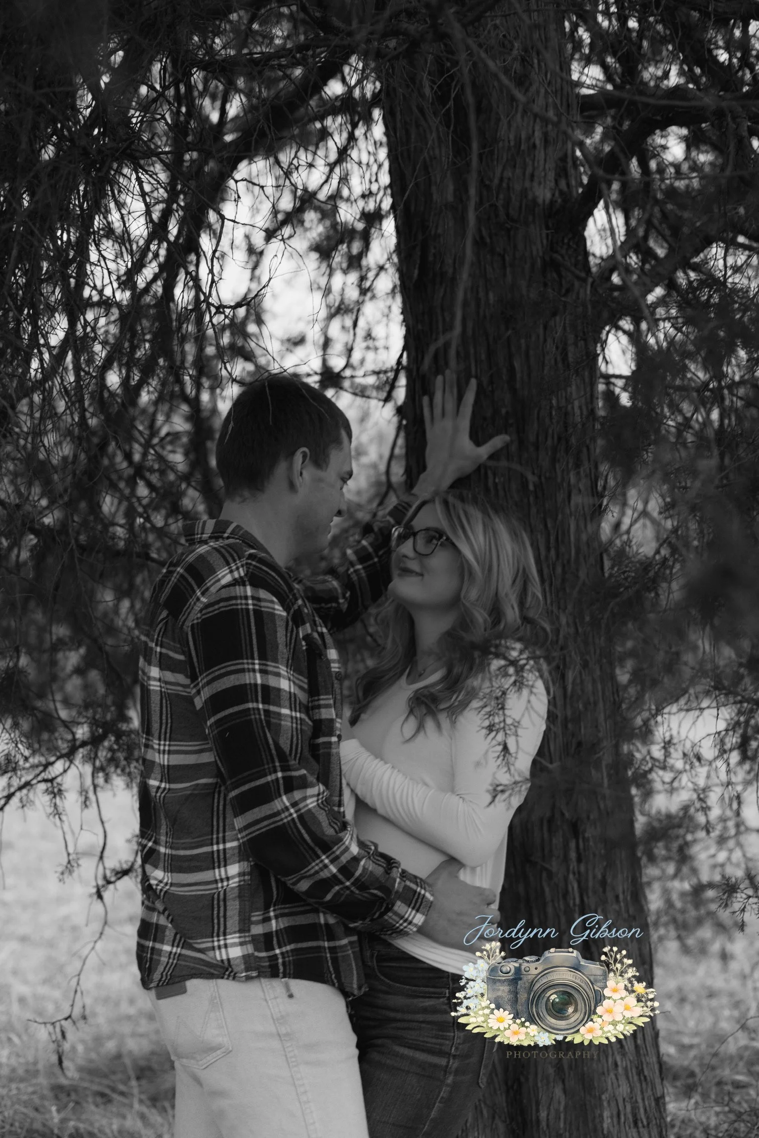A black and white photo of a couple standing close to each other outdoors next to a large tree. The man is touching the tree with one hand and looking at the woman, who is smiling and looking back at him. The woman is wearing glasses and a long-sleev