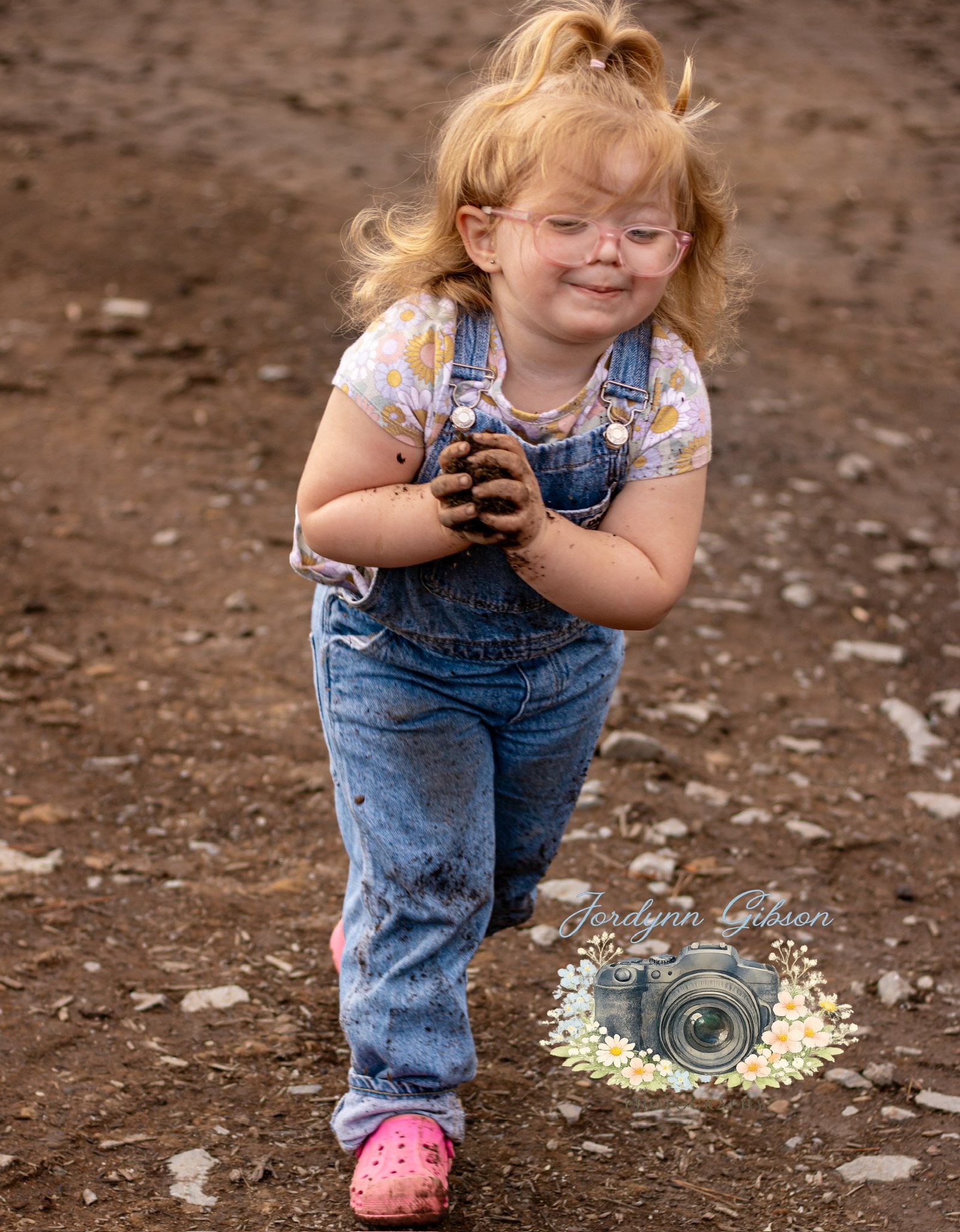 A young girl with curly red hair, wearing glasses, blue overalls, a floral t-shirt, and pink shoes, is running on dirt with a muddy face and hands, smiling as she plays outside.