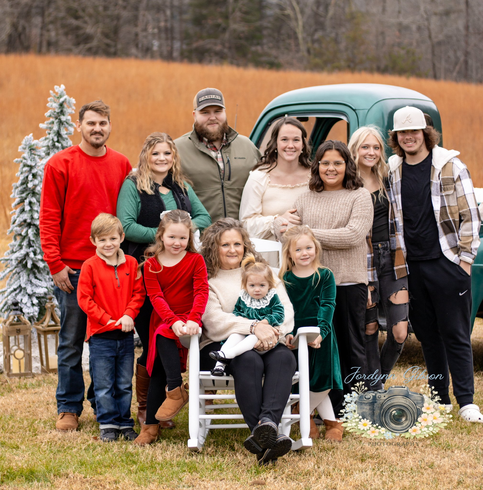Family outdoor portrait in fall colors with a vintage truck, some children and adults dressed casually, some in holiday colors, and a small decorated Christmas tree on the left.