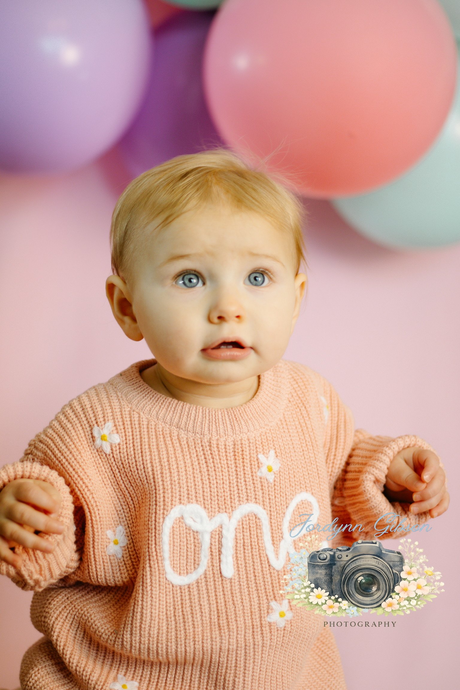 A young child with blonde hair and blue eyes, wearing a peach sweater with white flowers and the word 'one' on it, standing in front of pink, purple, and white balloons.