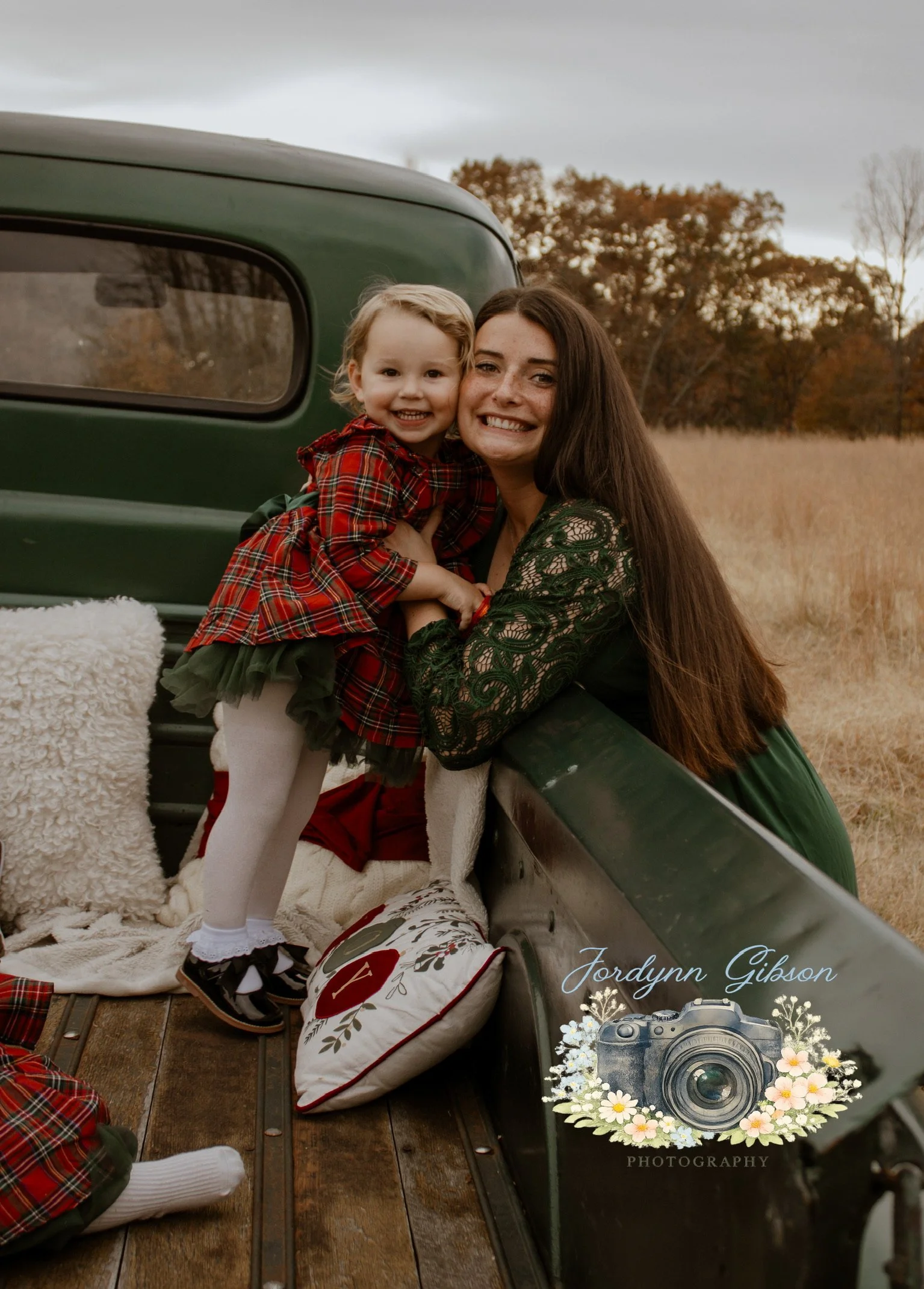 A woman and a young girl hugging and smiling in the back of a vintage green truck with fall foliage in the background.