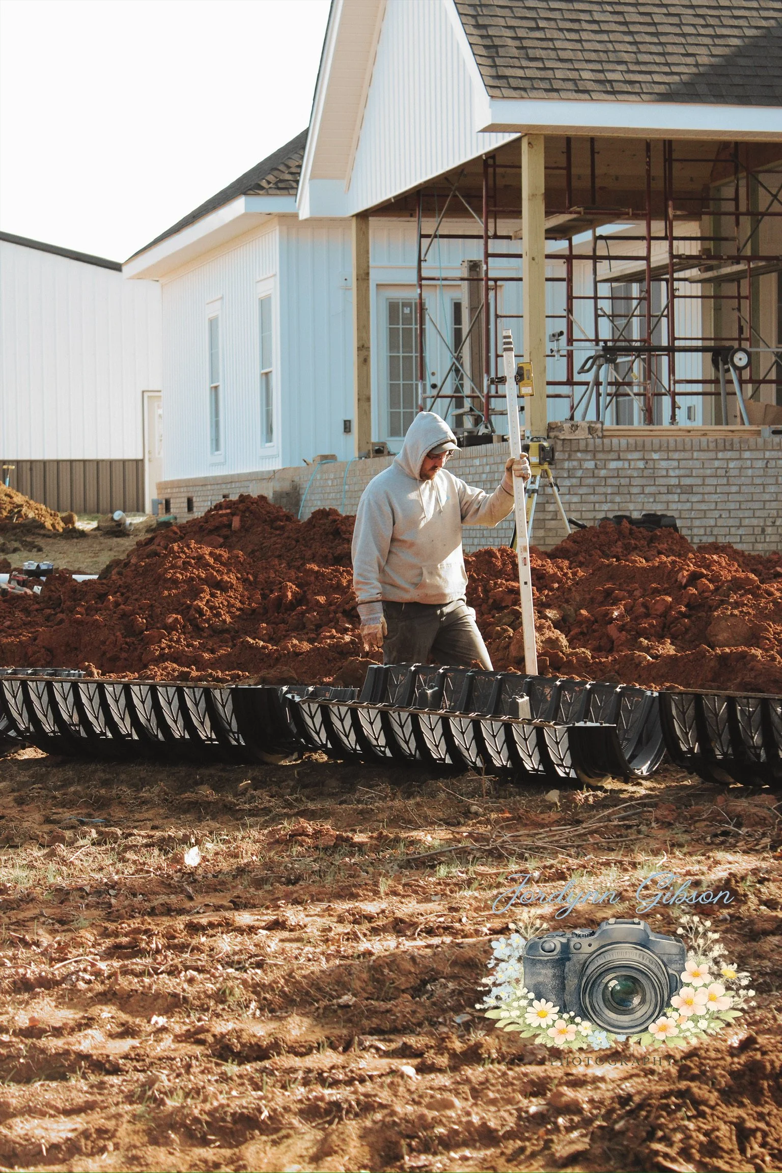 A construction worker on a dirt lot measuring a level in front of a house under construction with scaffolding.