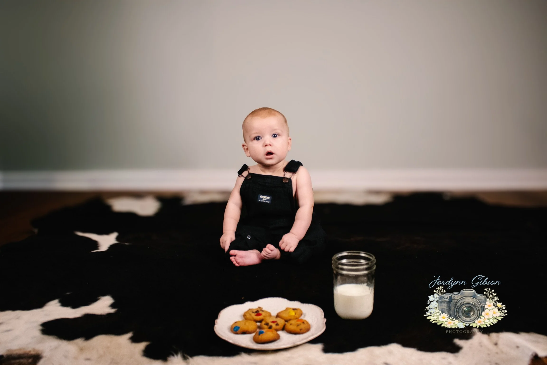 A baby sitting on a black and white cowhide rug in front of a plain wall, with a plate of cookies, a jar of milk, and a logo with a camera and flowers in the bottom right corner.