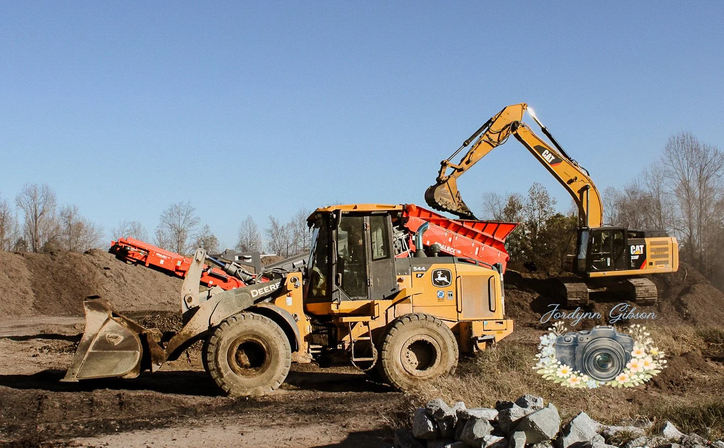 Construction site with a bulldozer and excavator working on dirt piles, in a rural area with trees in the background.