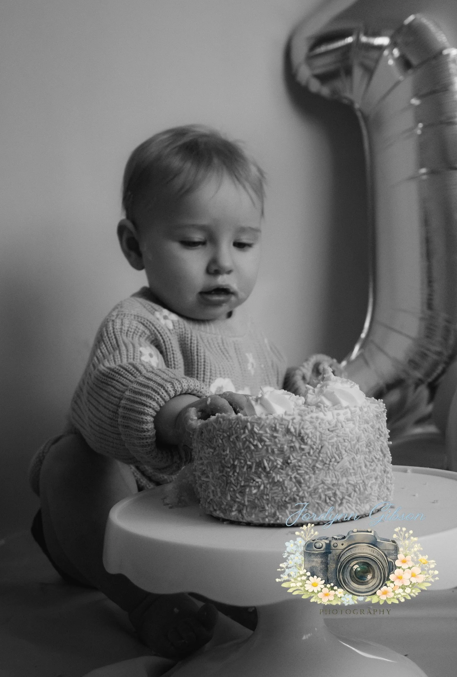 A young child with short hair looks at a birthday cake on a high chair tray. The cake is decorated with sprinkles and frosting. A balloon in the shape of the number 1 is in the background.