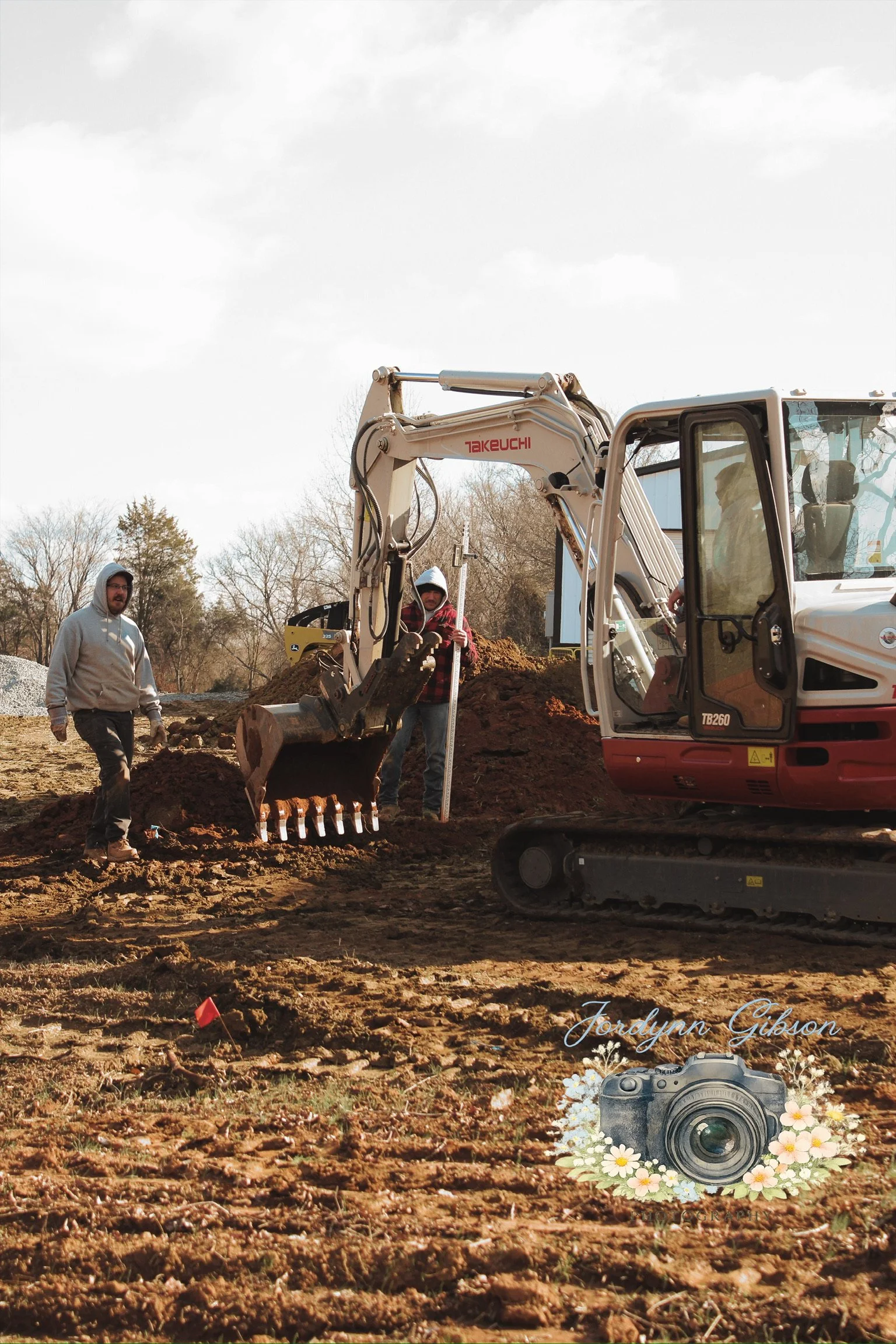 Two men working at a construction site with an excavator digging into the ground, surrounded by dirt and trees in the background, under a partly cloudy sky.