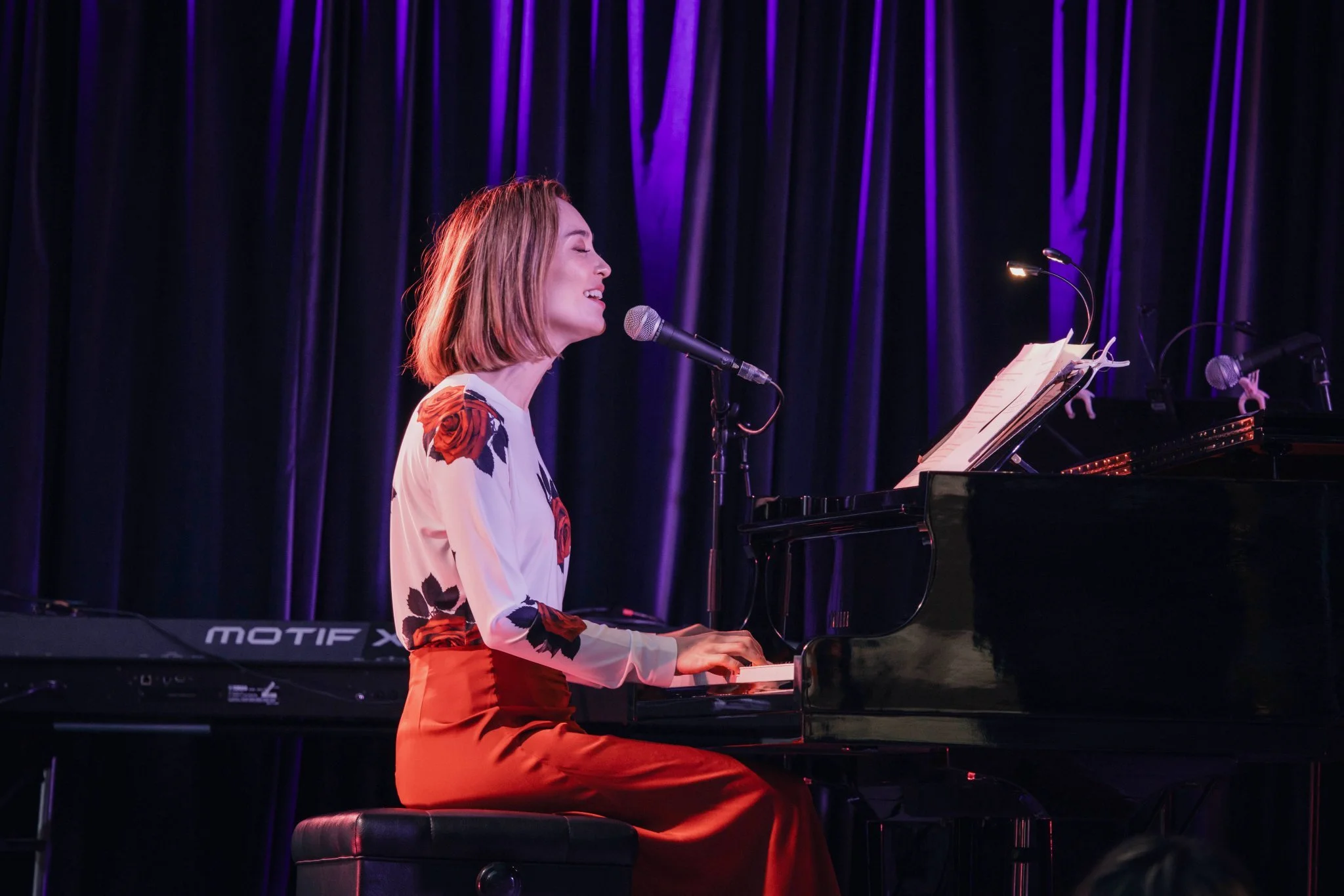 Woman singing and playing a grand piano on stage, with purple stage lights and black curtains in the background.