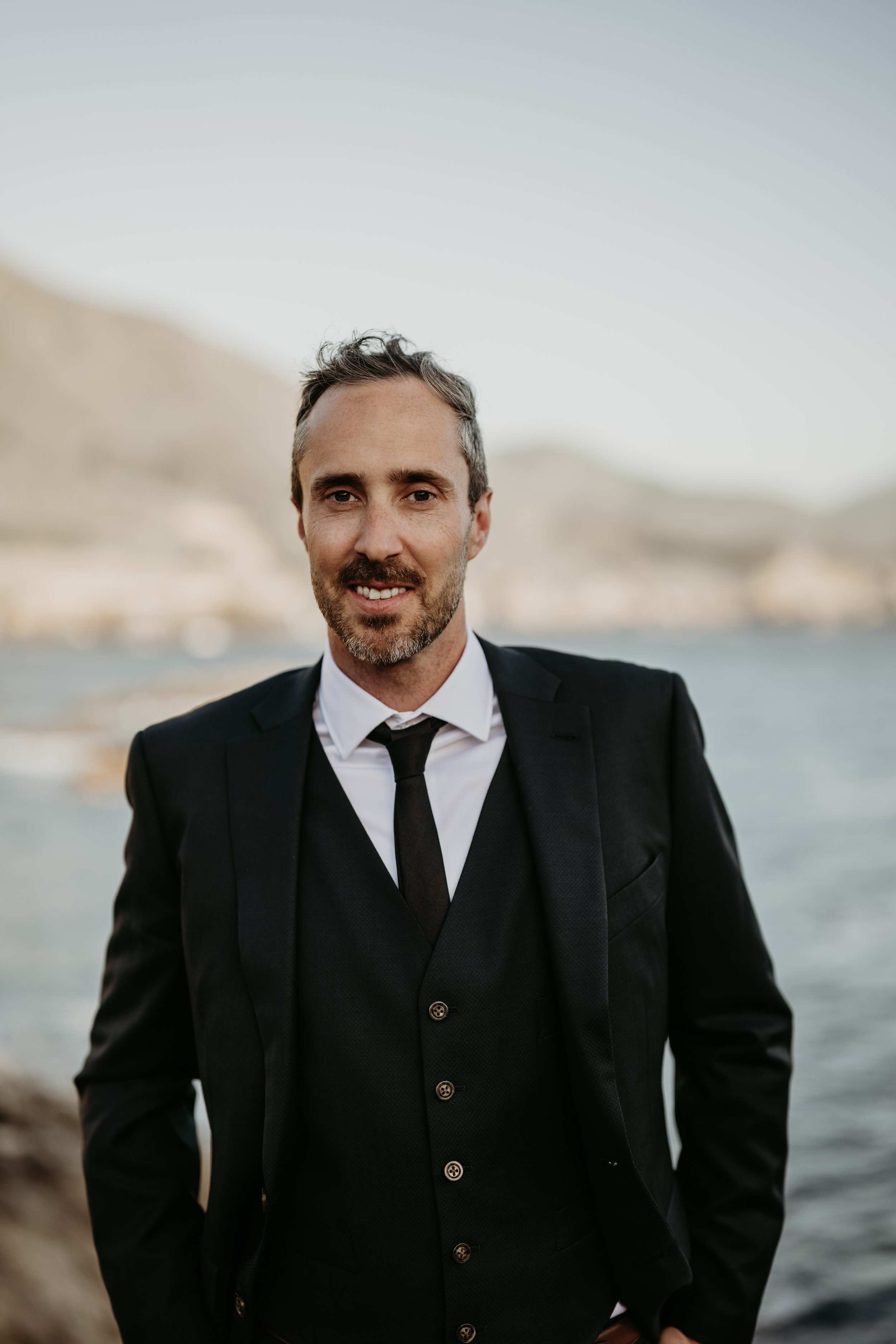 A man in a black suit and tie standing outdoors near the water with a blurred mountainous landscape in the background.