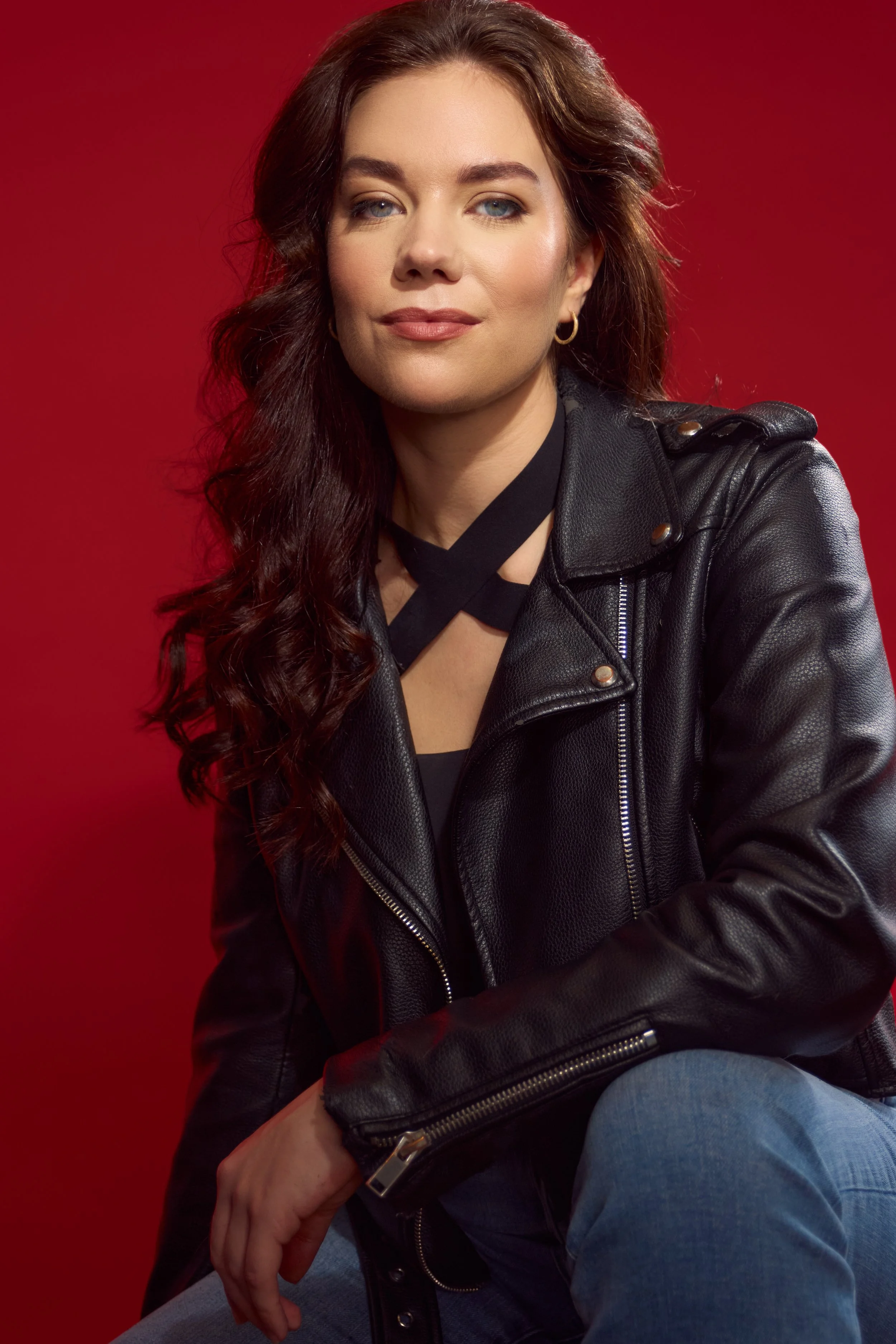 A woman with long wavy brown hair, blue eyes, and light makeup, wearing a black leather jacket and blue jeans, sitting against a red background.
